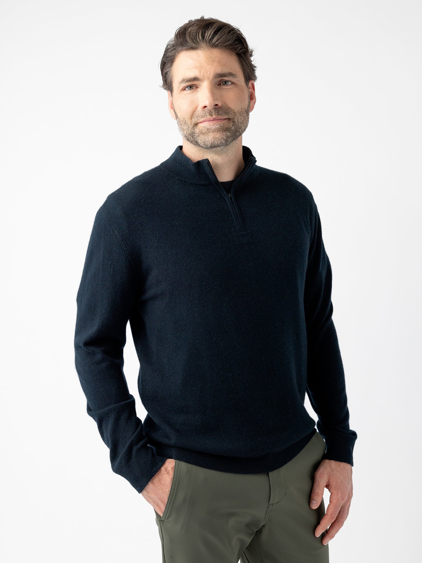 A man with short, dark hair and a beard is standing against a plain white background. He is wearing a Men's Quarter Zip Sweater by Cozy Earth, which is dark and long-sleeved, paired with olive-green pants. His hands are in his pockets, and he has a neutral expression on his face. 