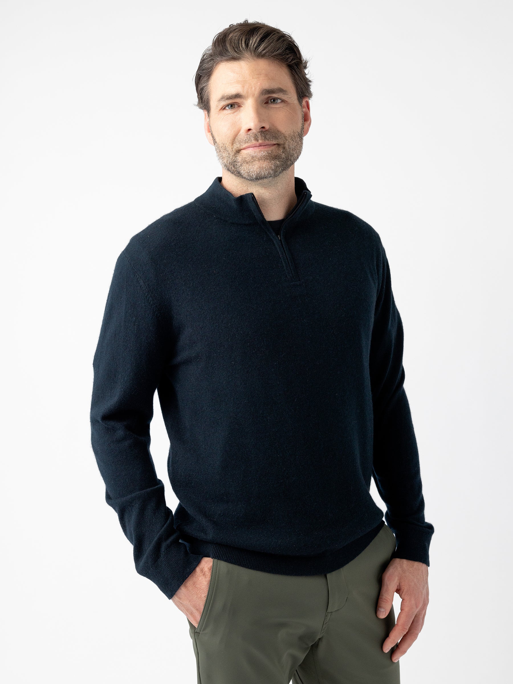 A man with short, dark hair and a beard is standing against a plain white background. He is wearing a Men's Quarter Zip Sweater by Cozy Earth, which is dark and long-sleeved, paired with olive-green pants. His hands are in his pockets, and he has a neutral expression on his face. |Color:Jet Black