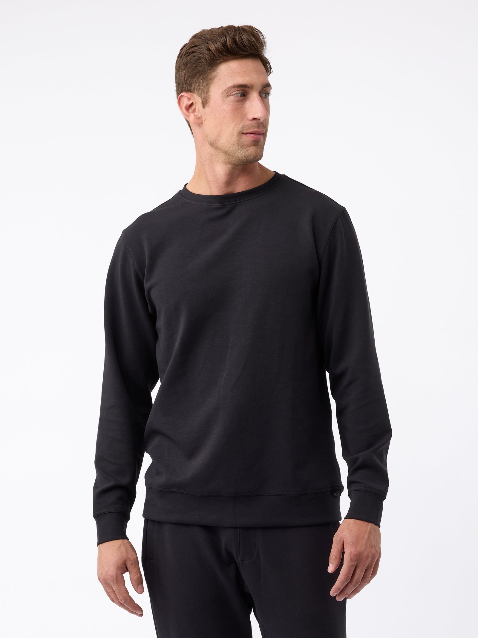 A man with short brown hair, dressed in a Cozy Earth Men's StretchTech Crewneck and black pants, stands against a plain white background. He is looking slightly to his right, with his hands relaxed at his sides. 