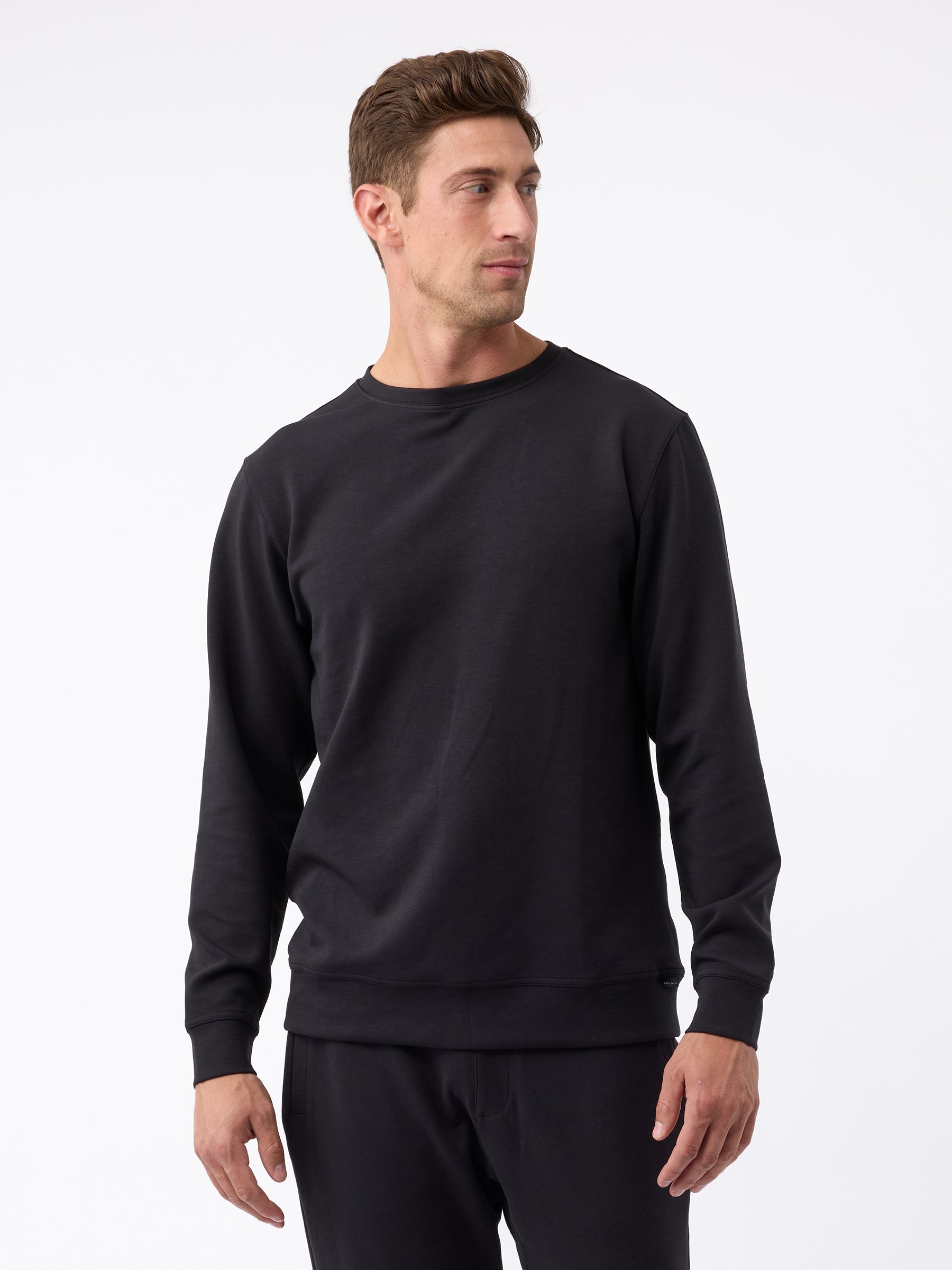 A man with short brown hair, dressed in a Cozy Earth Men's StretchTech Crewneck and black pants, stands against a plain white background. He is looking slightly to his right, with his hands relaxed at his sides. |Color:Jet Black