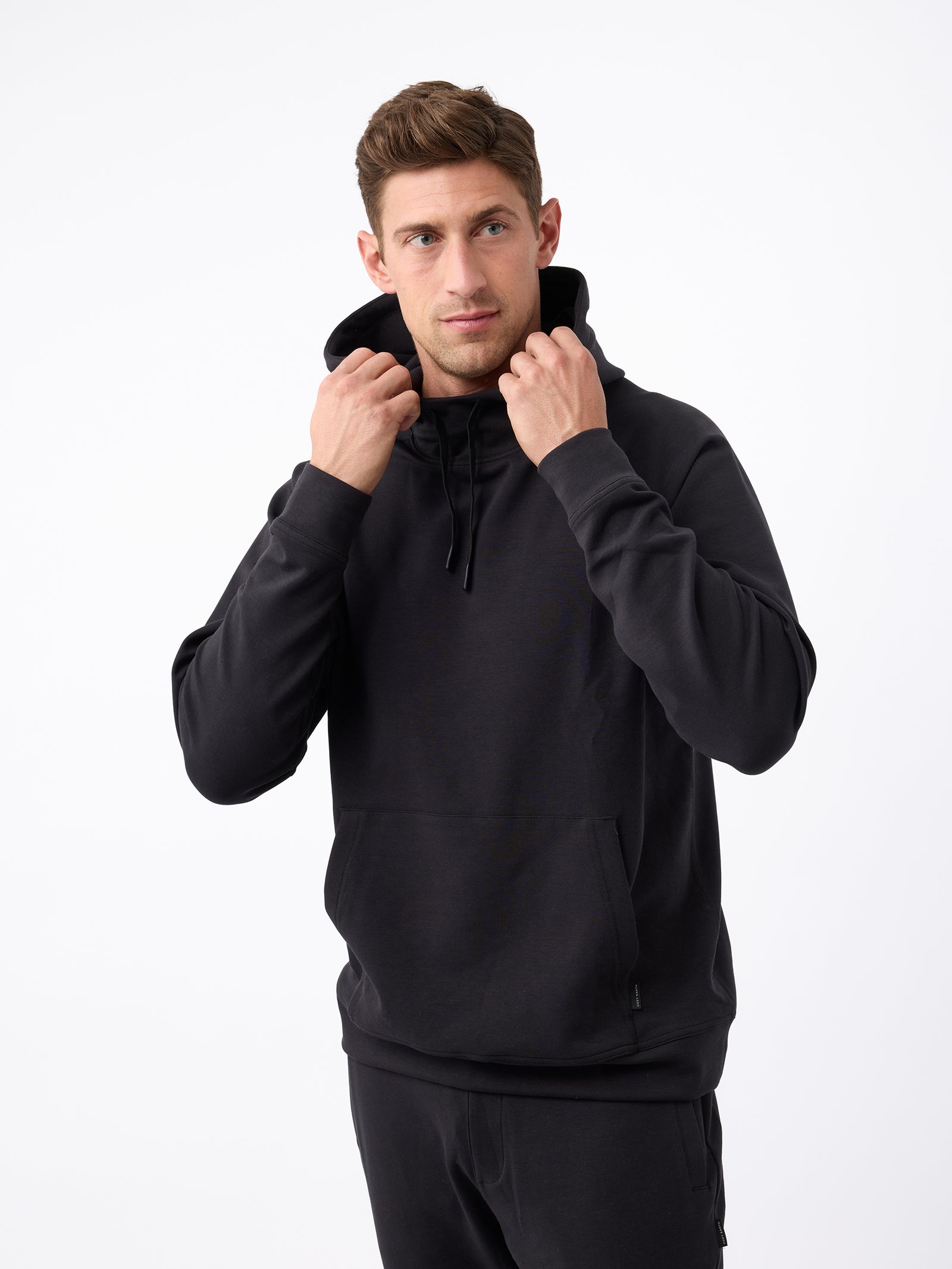 A man standing against a plain white background is wearing a black Men's StretchTech Hoodie and black pants from Cozy Earth. He is holding the hoodie strings with both hands. The man has short brown hair and a serious expression. 