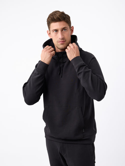 A man standing against a plain white background is wearing a black Men's StretchTech Hoodie and black pants from Cozy Earth. He is holding the hoodie strings with both hands. The man has short brown hair and a serious expression. |Color:Jet Black