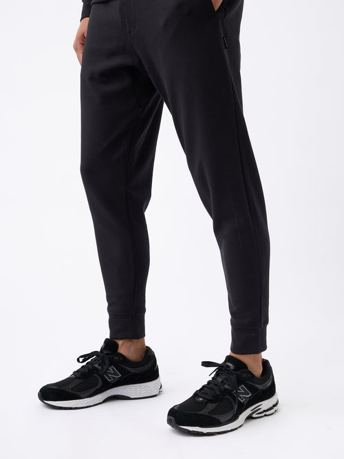 A person is wearing Cozy Earth's Men's StretchTech Jogger and black New Balance running shoes with white soles. The person’s hands are in their pockets, and only the lower half of their body is visible against a plain white background. |Color:Jet Black