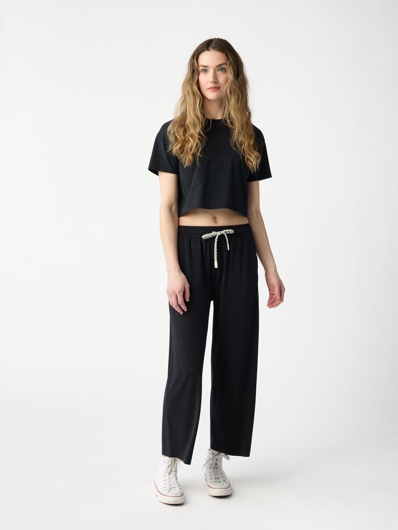 A person with long wavy hair stands against a plain background, wearing a black crop top, Cozy Earth's Women's Studio Cropped Wide Leg Pant in black, and white sneakers. Their expression is neutral, and their arms are relaxed by their sides. |Color:Jet Black