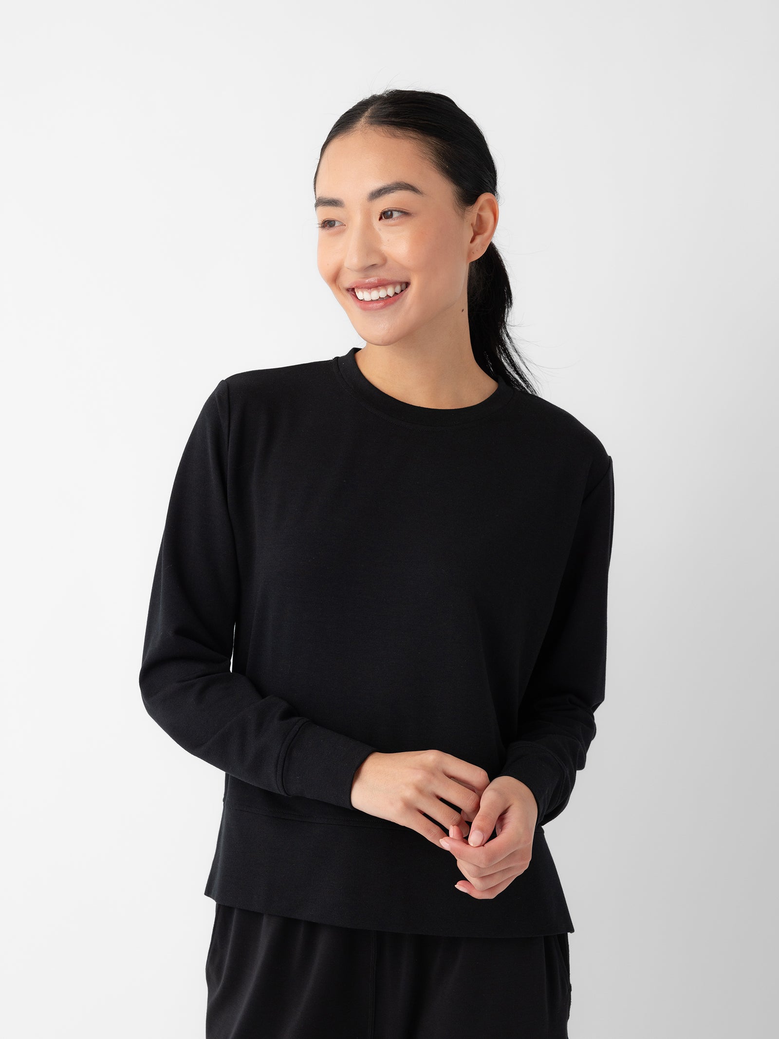 A person with long black hair tied back, wearing a Cozy Earth Women's StudioLite Crewneck in plain black, stands against a white background. They are smiling and looking slightly to the side while loosely clasping their hands at their waist. 