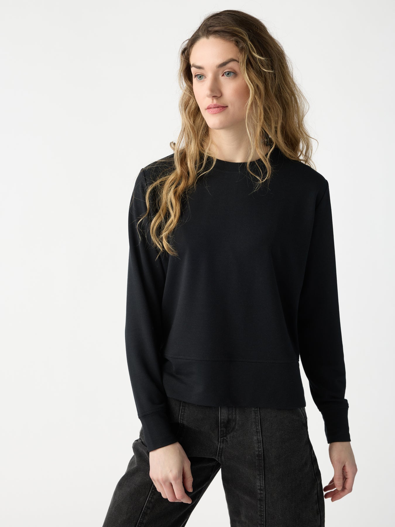 A woman with long wavy hair poses in a Cozy Earth Women's StudioLite Crewneck and dark jeans, with a neutral expression gazing slightly to the side against a simple white backdrop. |Color:Jet Black