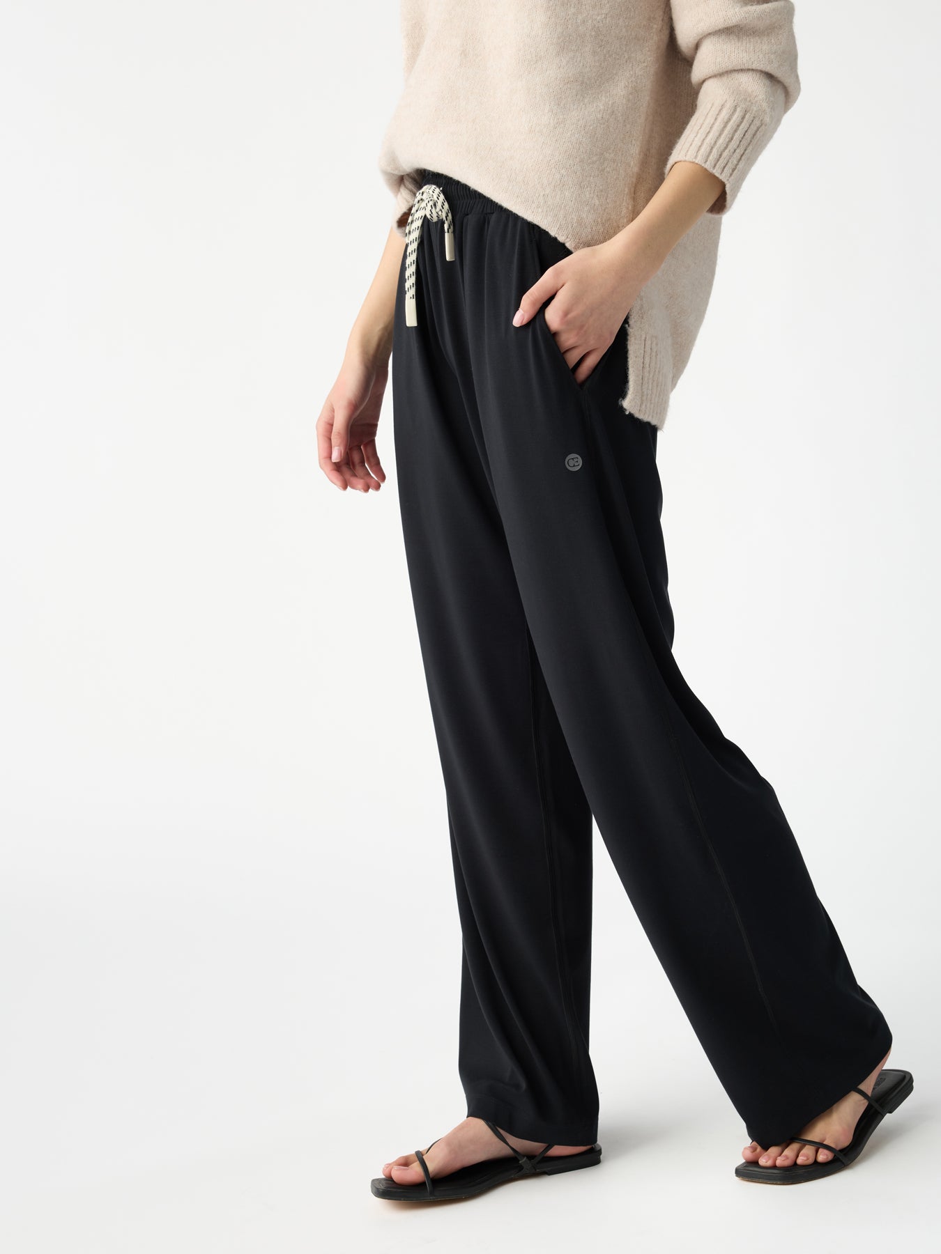 A person models a relaxed, casual look wearing Cozy Earth's Women's Studio Wide Leg Pant in black and a beige sweater paired with matching black sandals against a plain background. |Color:Jet Black