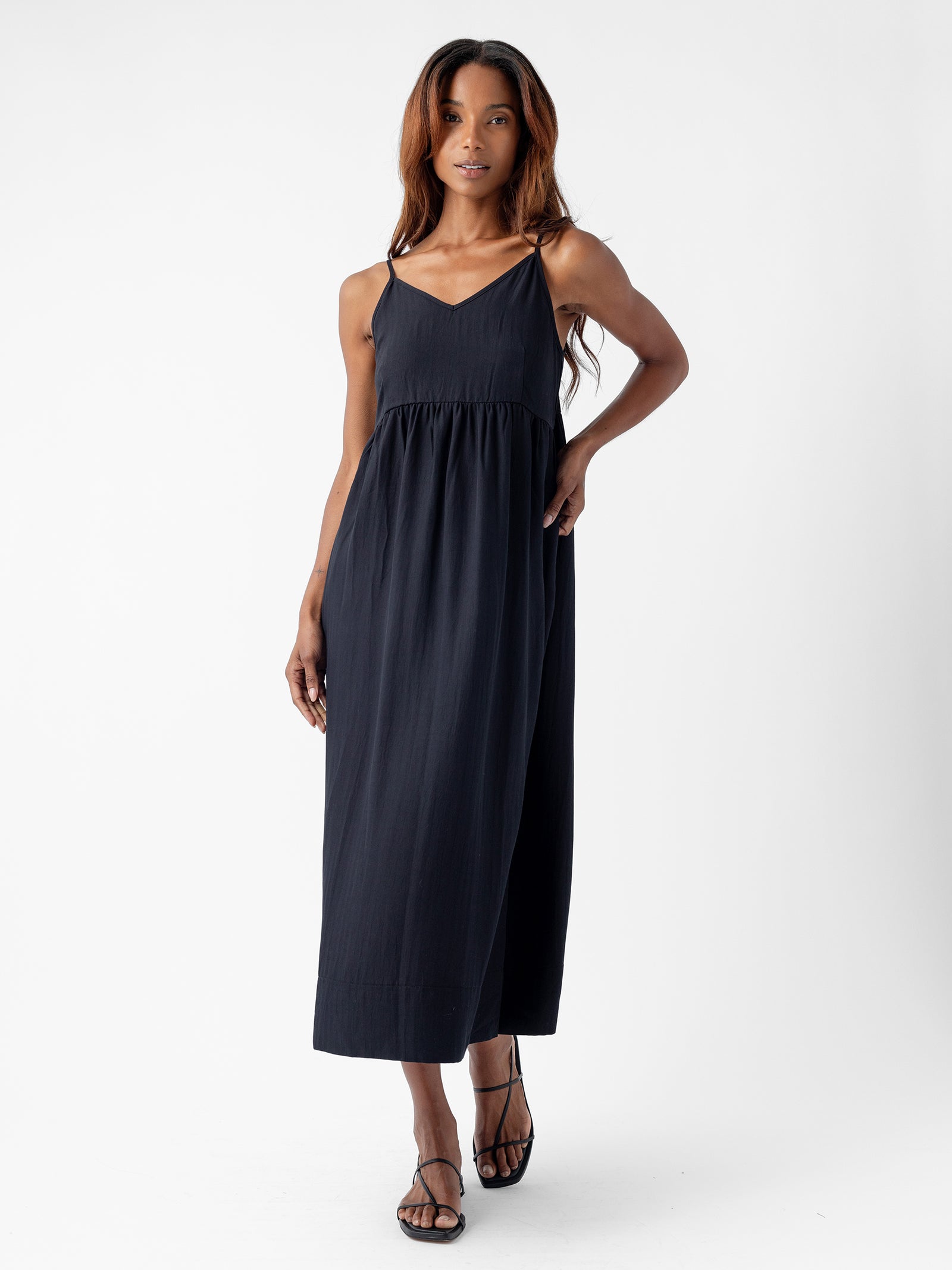 A woman with long hair is standing against a plain background, wearing Cozy Earth's Women's Sunset Gathered Dress, which is a sleeveless, ankle-length black dress with thin straps and a slightly gathered waist. She is also wearing black sandals and has one hand placed on her hip. 