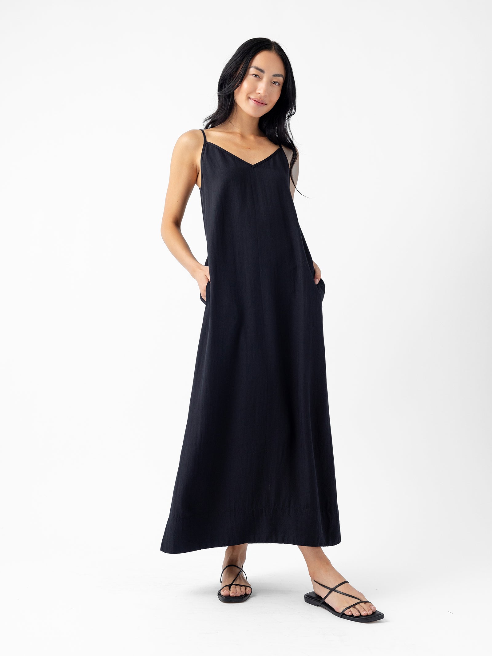 A person with long, dark hair wearing the Cozy Earth Women's Sunset Slip Dress stands against a plain white background. They wear black strappy sandals and have their hands in the dress's side pockets, smiling gently. 