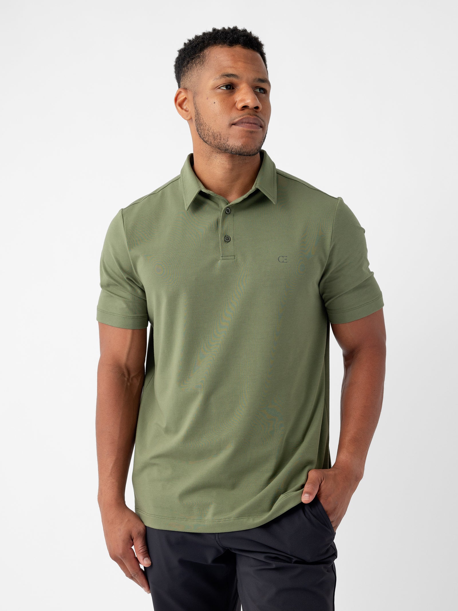 A person stands against a plain background wearing the Men's Everyday Polo by Cozy Earth in green, paired with black pants. They have short hair and are looking off to the side with one hand in their pocket. 