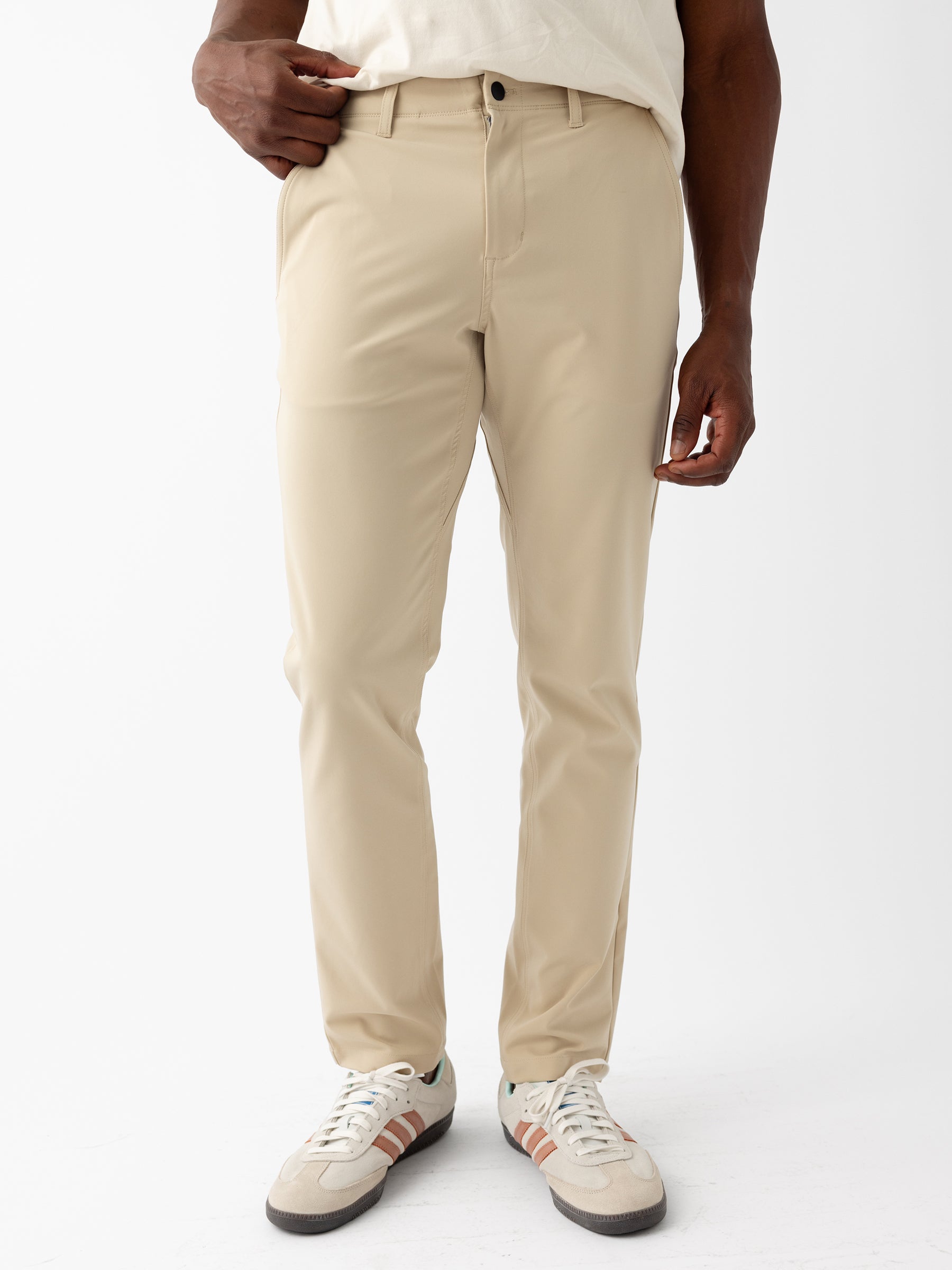A person wearing Cozy Earth's Men's Everywhere Pant 30L in beige and a white shirt stands with one hand in their pocket. They are also wearing white sneakers featuring beige and green accents. The background is plain white. |Color:Khaki