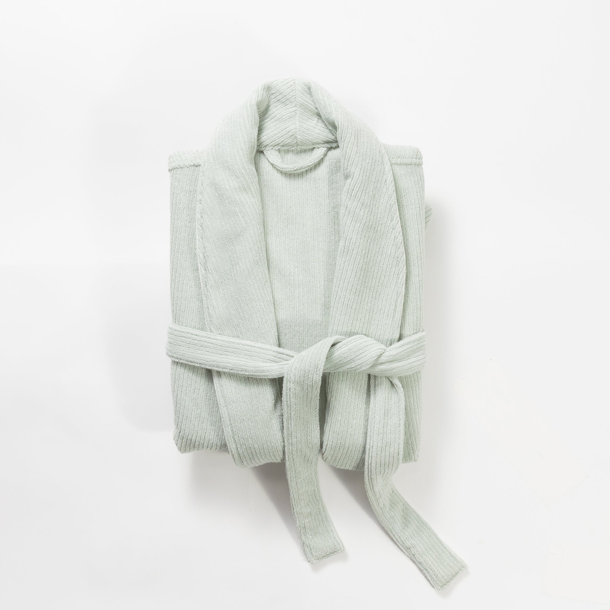 The Ribbed Terry Bath Robe by Cozy Earth is neatly folded and tied with its belt around the middle, creating a tidy, compact appearance. The light gray robe features a soft, textured fabric and a shawl collar, all presented against a plain white background. |Color:Laurel