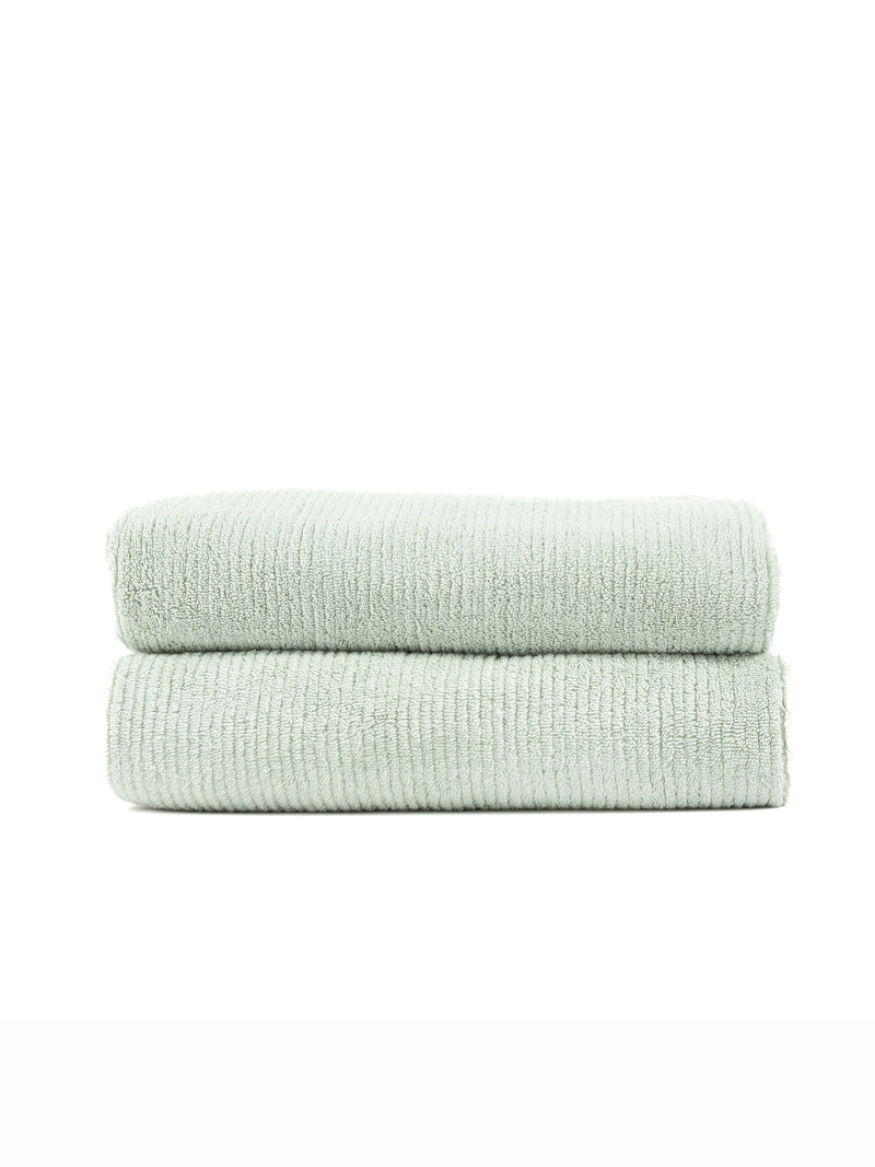 Two Ribbed Terry Bath Towels by Cozy Earth, in light green, are neatly folded and stacked together against a plain white background.