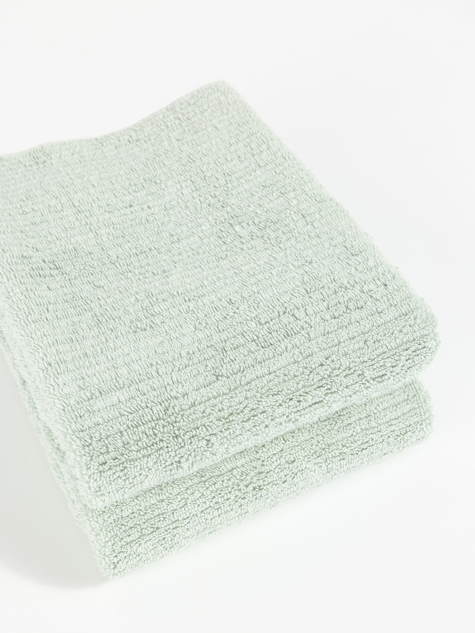 Two neatly folded Ribbed Terry Hand Towels by Cozy Earth in light green are stacked on top of each other against a plain white background. The towels appear soft and fluffy with a simple, textured pattern. 