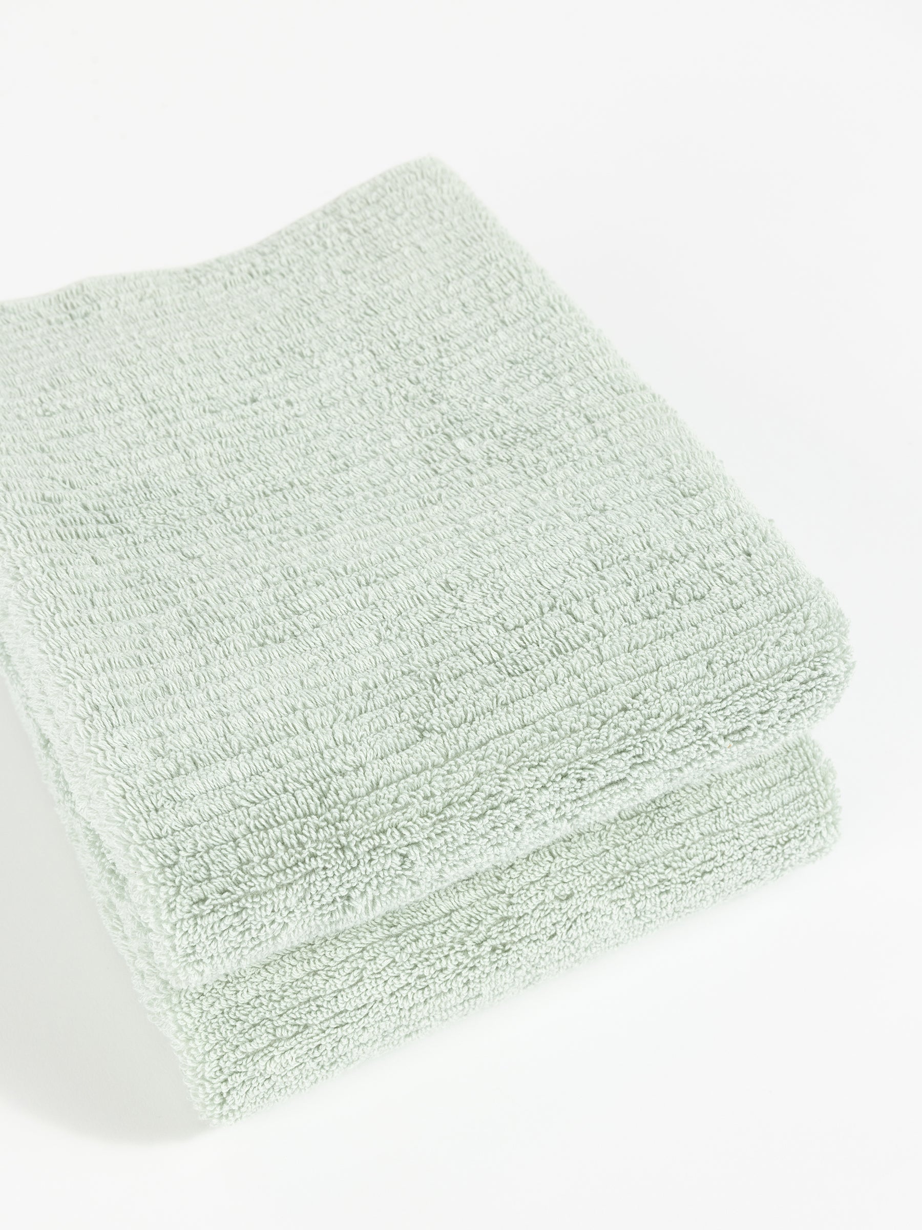 Two neatly folded Ribbed Terry Hand Towels by Cozy Earth in light green are stacked on top of each other against a plain white background. The towels appear soft and fluffy with a simple, textured pattern. |Color:Laurel