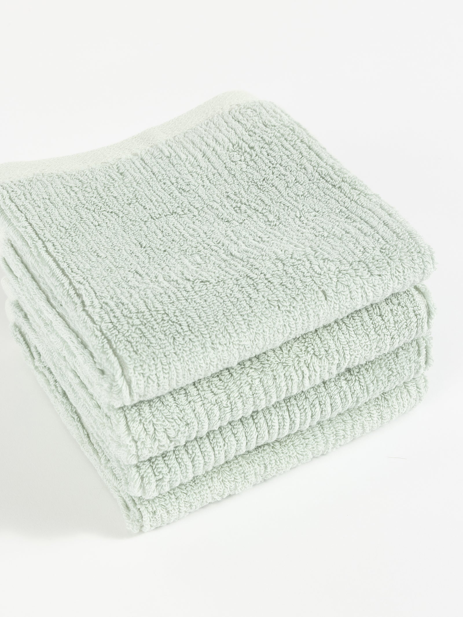 A neatly folded stack of four soft, light green Ribbed Terry Washcloths by Cozy Earth is placed on a white surface. The washcloths have a textured appearance and appear fluffy, suggesting they are absorbent and comfortable for use. 