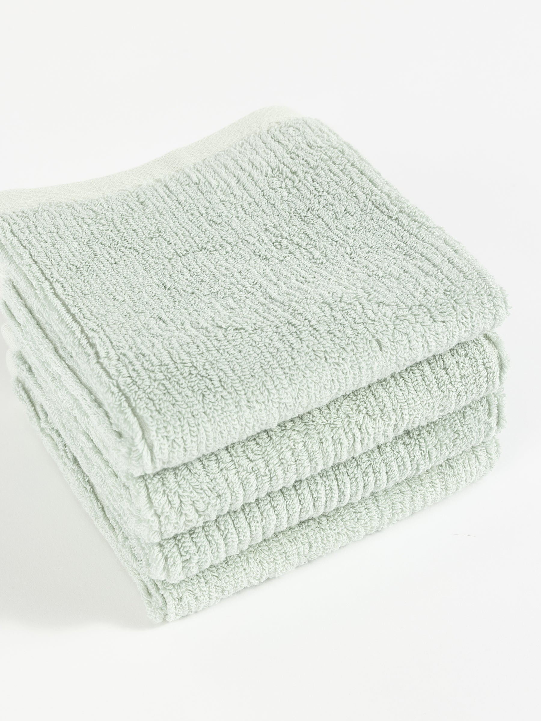 A neatly folded stack of four soft, light green Ribbed Terry Washcloths by Cozy Earth is placed on a white surface. The washcloths have a textured appearance and appear fluffy, suggesting they are absorbent and comfortable for use. |Color:Laurel