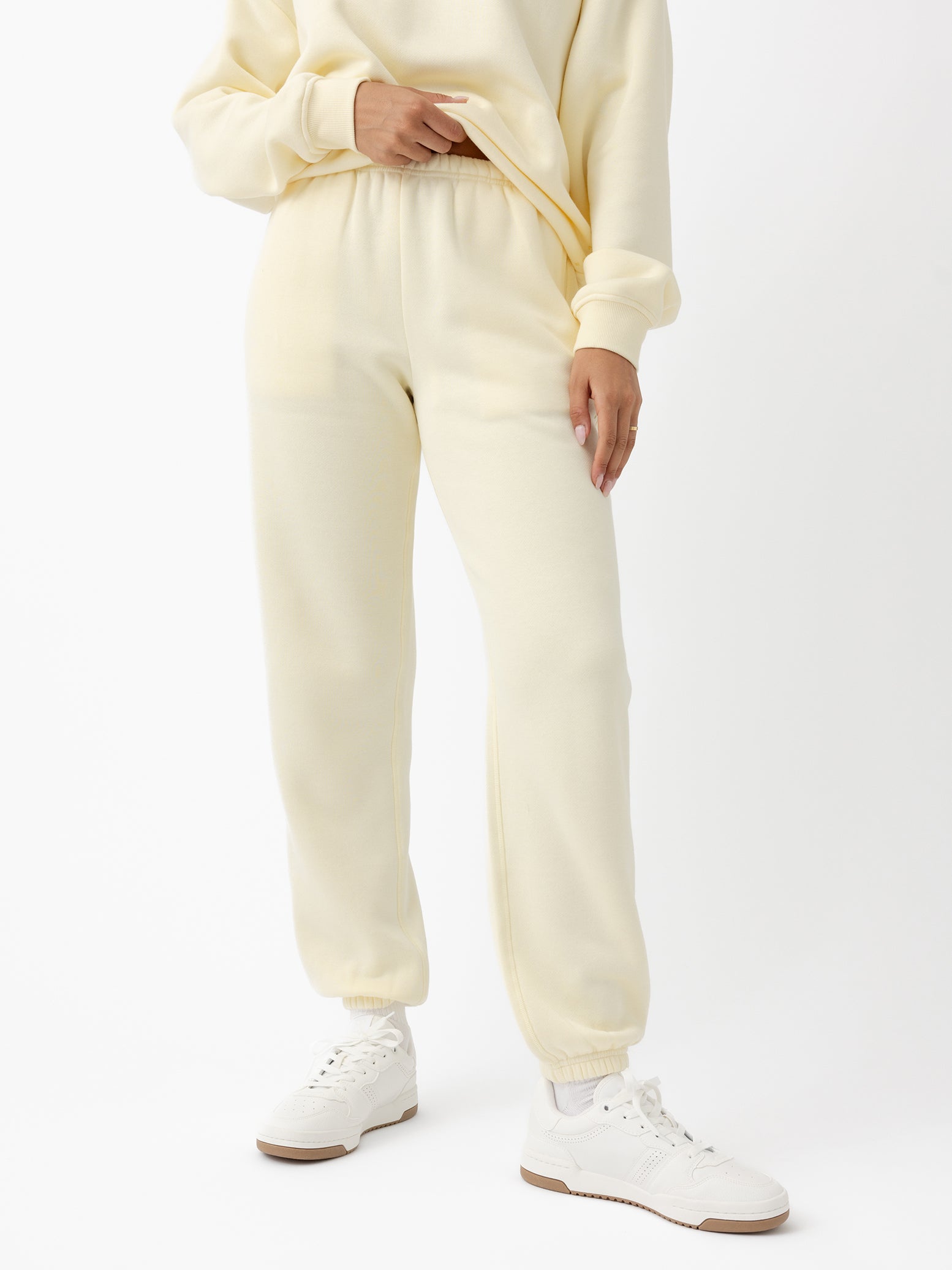 A person is dressed in Cozy Earth's Women's CityScape Sweatpant, a cream-colored sweatshirt, and white sneakers with brown soles, holding the sweatshirt near the waist against a plain white background. |Color:Lemonade