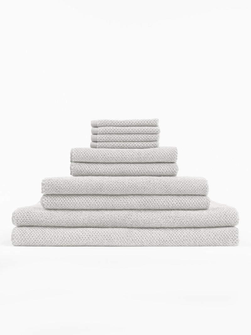 The Cozy Earth Complete Nantucket Bath Bundle features clean, plush light gray towels in various sizes with a textured pattern, neatly stacked in a pyramid shape against a white background.