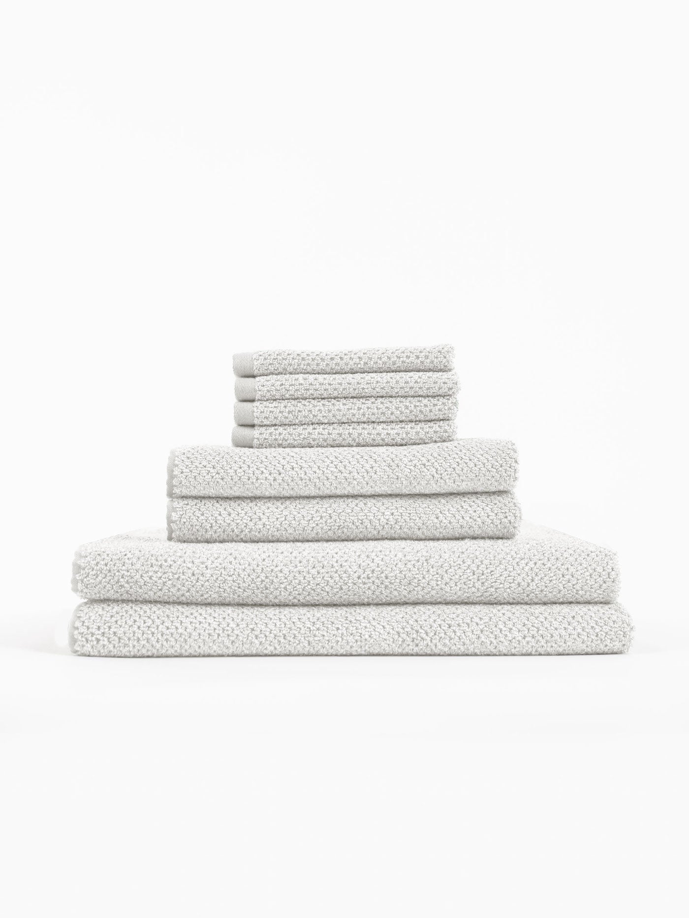The Cozy Earth Nantucket Bath Towel Set features textured, light gray towels in assorted sizes, neatly stacked on a white background.