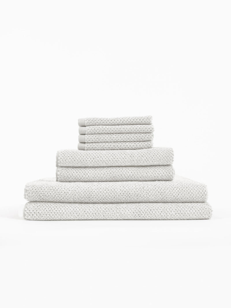 The Cozy Earth Nantucket Bath Towel Set features textured, light gray towels in assorted sizes, neatly stacked on a white background.