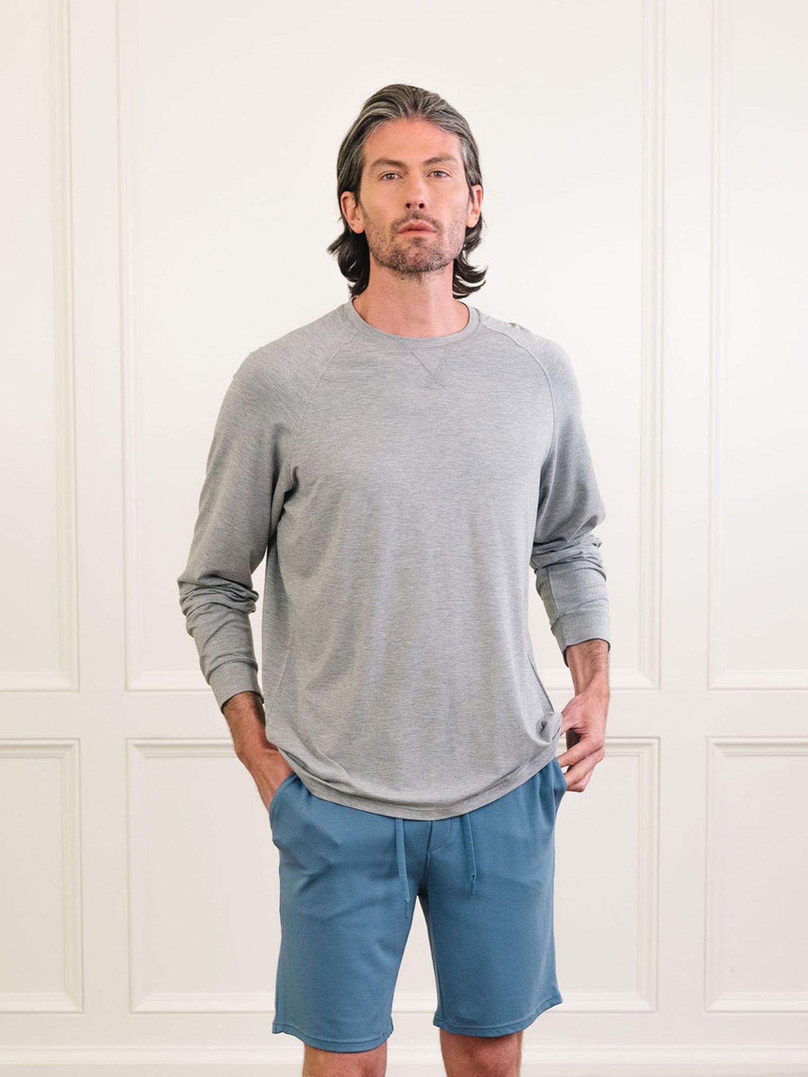 A man with shoulder-length hair stands facing forward against a neutral background. He is wearing the light gray Men's Stretch-Knit Long Sleeve Bamboo Pajama Set from Cozy Earth, paired with blue shorts, and has his hands in his pockets. The setting appears to be indoors with white paneled walls. 