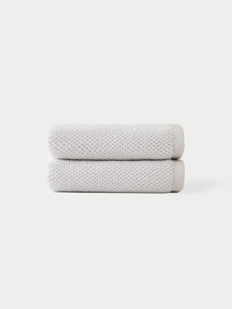 Two Nantucket Bath Towels by Cozy Earth, in light gray, are neatly folded and stacked against a plain white background.