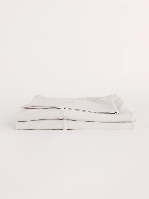 A neatly folded Bamboo Jersey Flat Sheet by Cozy Earth is displayed against a white background. The light gray sheets are tightly stacked, showcasing their smooth texture and clean appearance. |Color:Light Grey