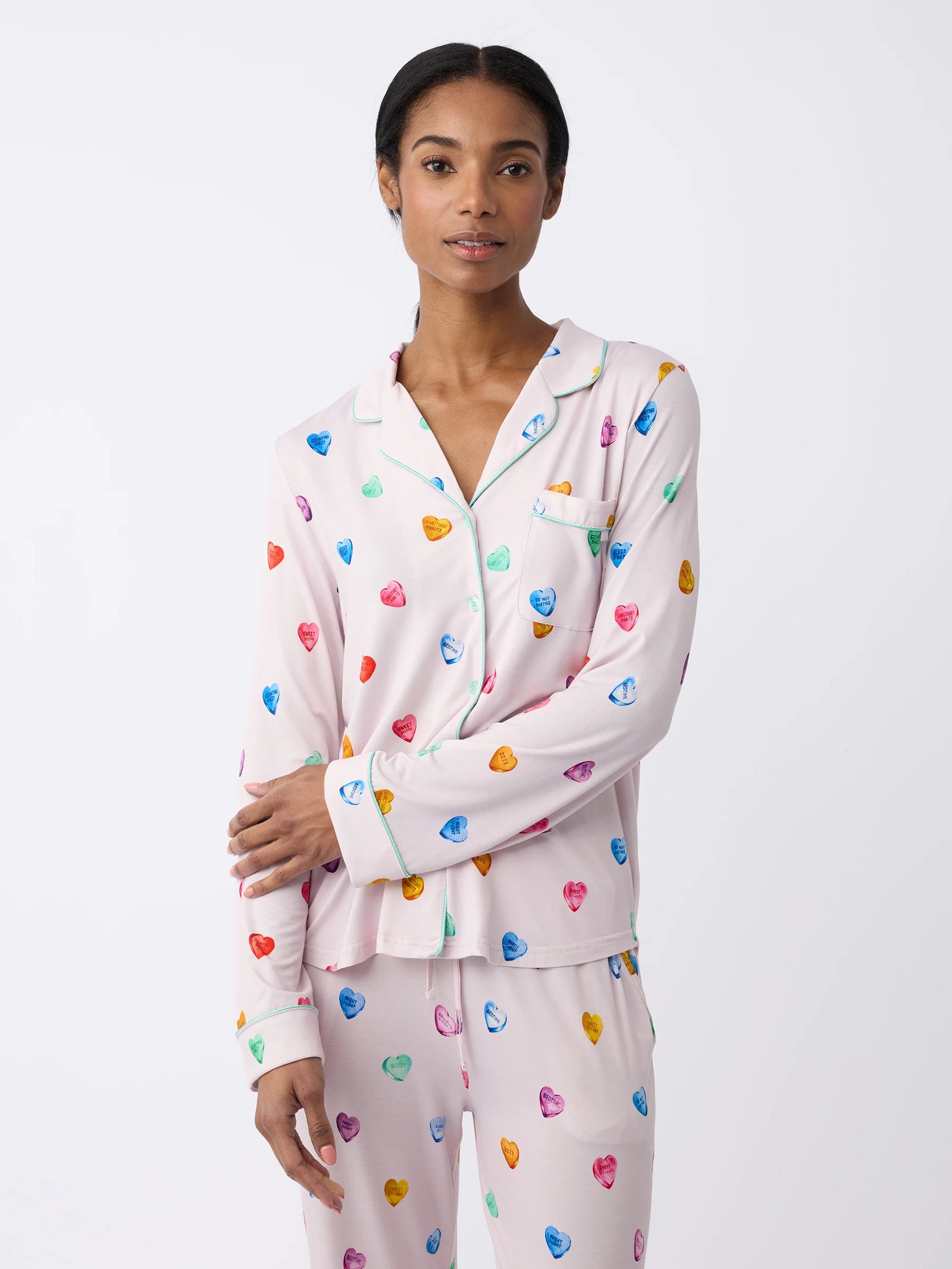 A person poses against a plain background wearing Cozy Earth's Women's Long Sleeve Bamboo Pajama Top in Stretch-Knit, featuring a light purple design with colorful heart patterns, a collar, and a front pocket. 