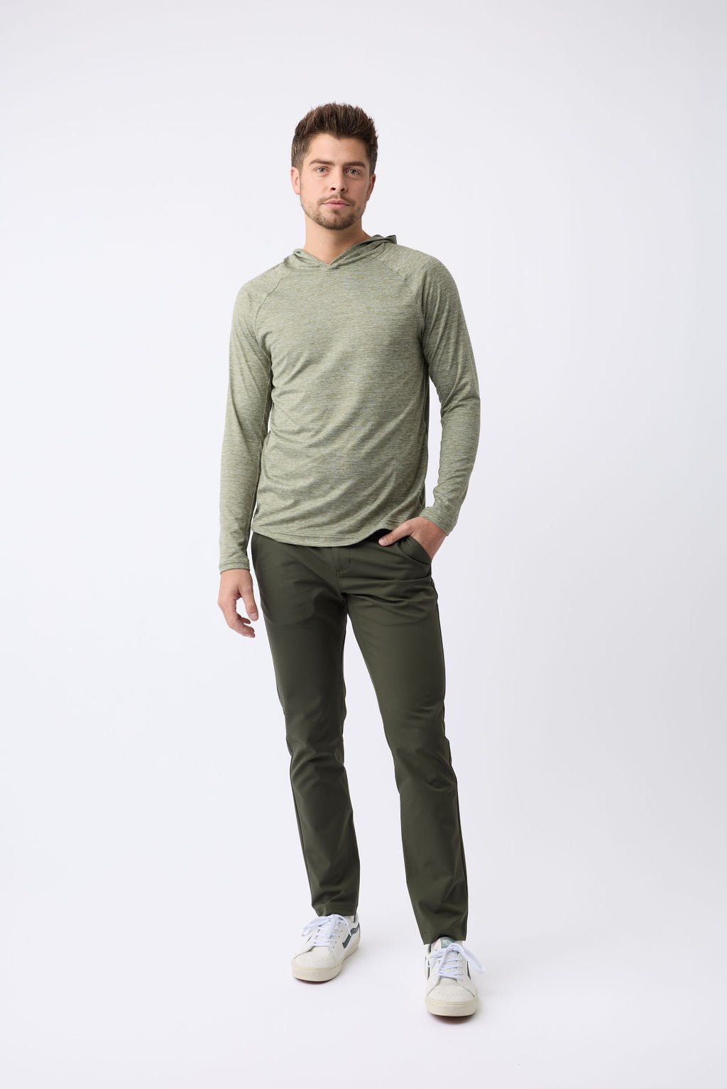A man stands against a plain light background wearing Cozy Earth's Men's Lightweight Performance Hoodie in light green paired with dark green pants. He has short hair and wears white sneakers, one hand in his pocket, the other by his side. 