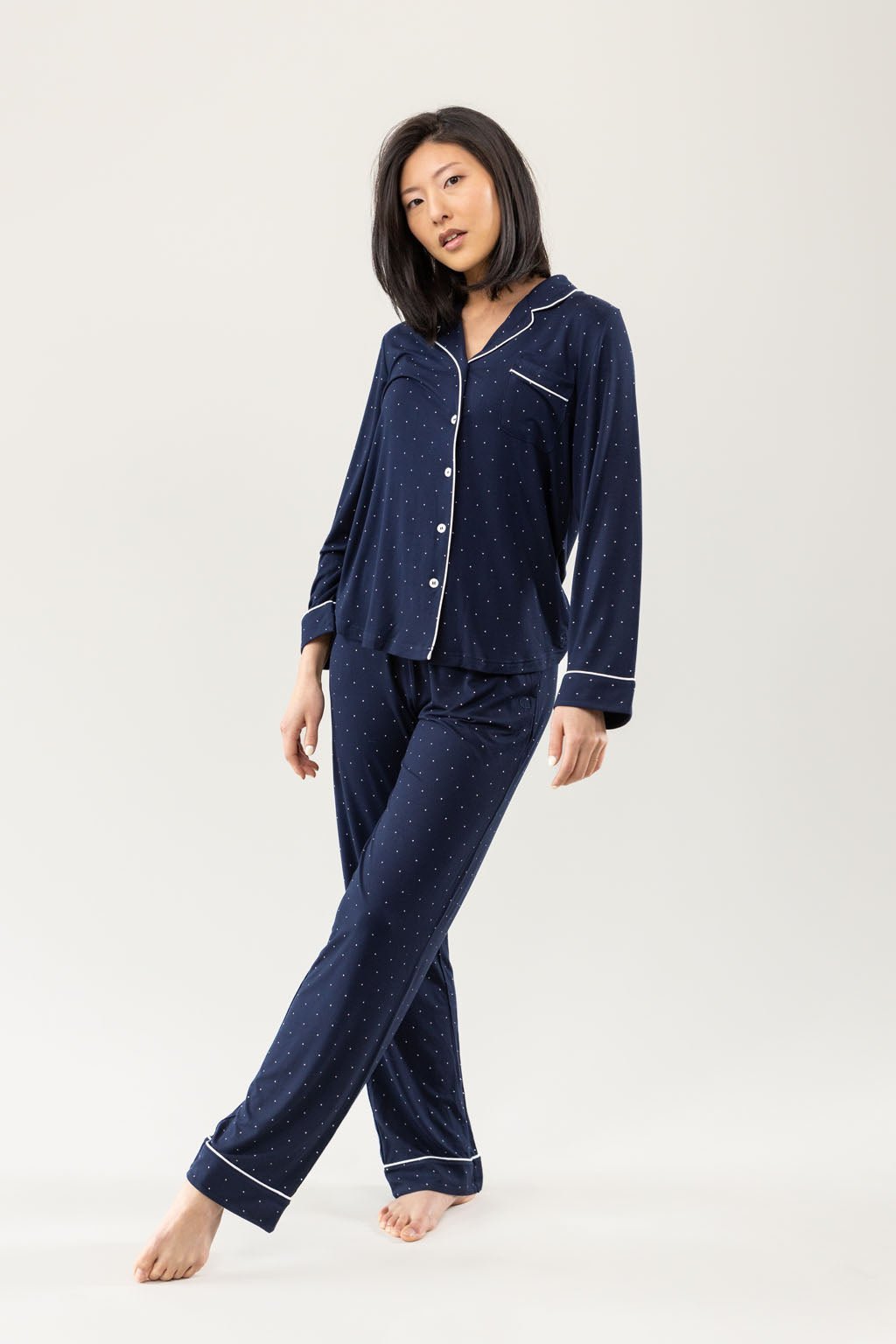 A person is barefoot in Cozy Earth's Women's Bamboo Stretch-Knit Long Sleeve Pajama Set, featuring navy blue fabric with white polka dots and piping. The set includes a button-up top and matching pants against a plain, light-colored background. 