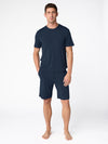 A barefoot man stands against a plain white background wearing the Cozy Earth Men's Bamboo Stretch-Knit Short Sleeve Pajama Set, with his hands in his pockets. |Color:Navy