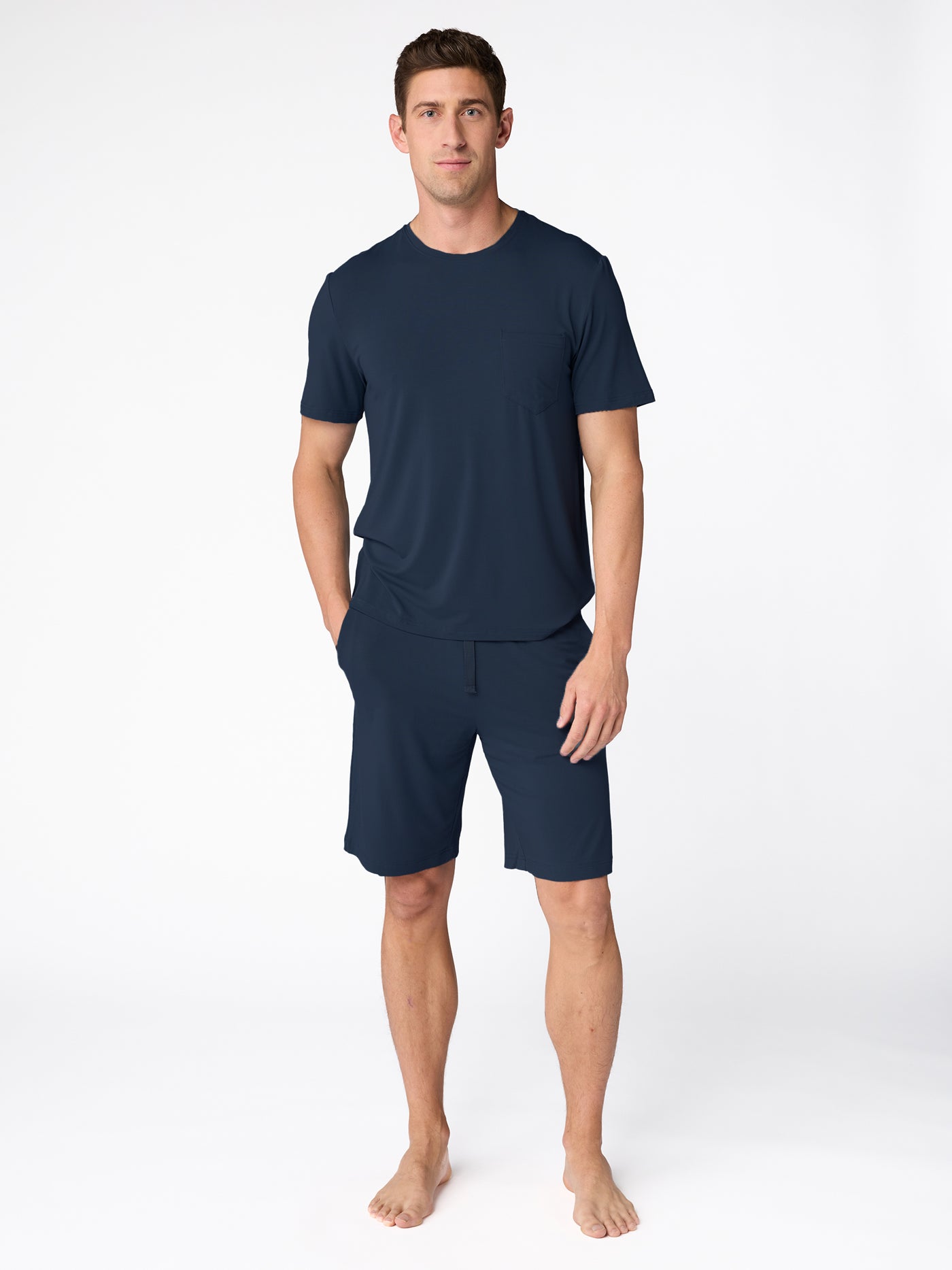A barefoot man stands against a plain white background wearing the Cozy Earth Men's Bamboo Stretch-Knit Short Sleeve Pajama Set, with his hands in his pockets. |Color:Navy