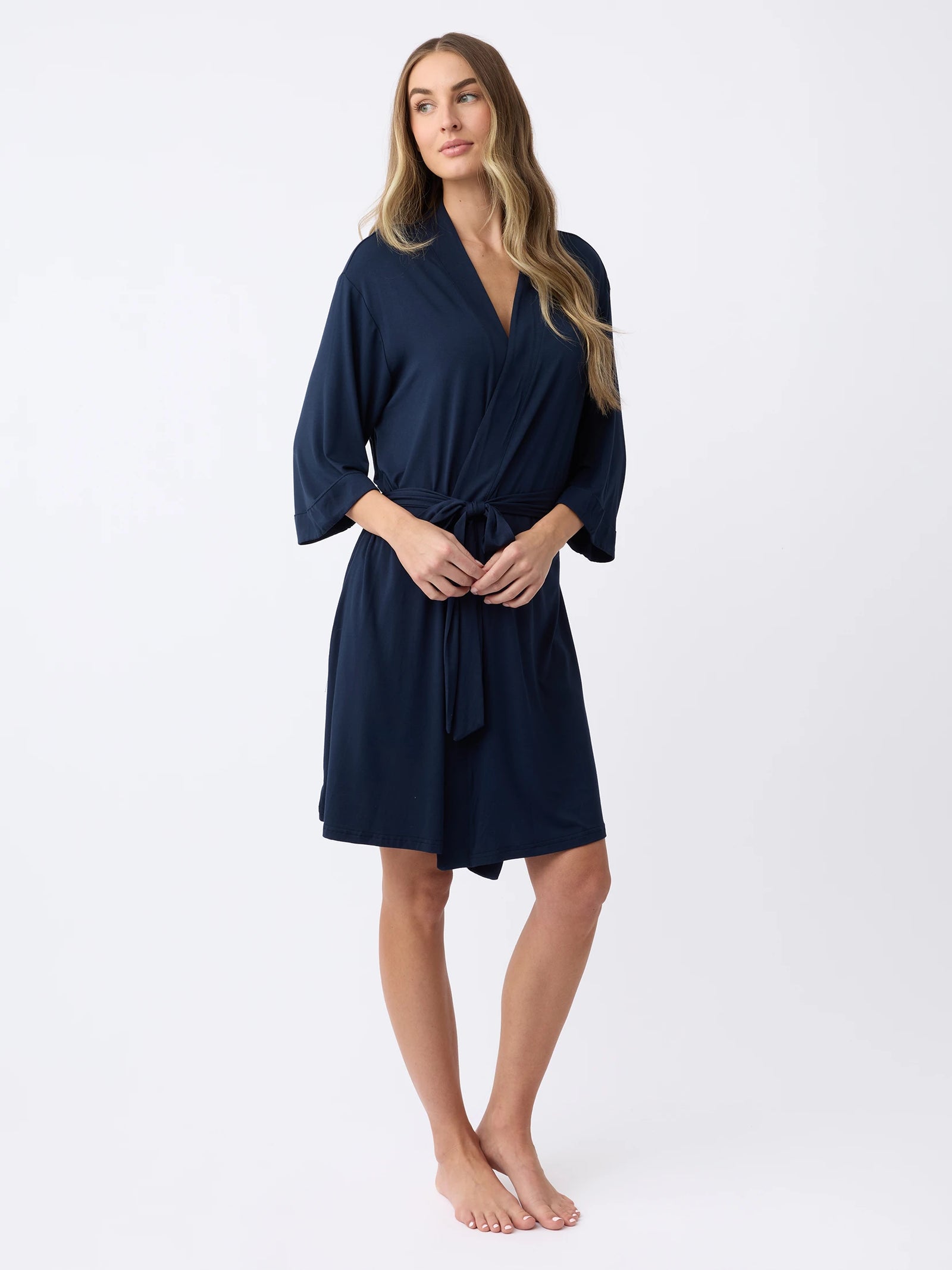 A woman stands barefoot against a plain white background, wearing a Cozy Earth Women's Stretch-Knit Bamboo Kimono Robe in dark blue. Her long, wavy hair complements her relaxed expression as she rests her hands on the robe's waist tie. 