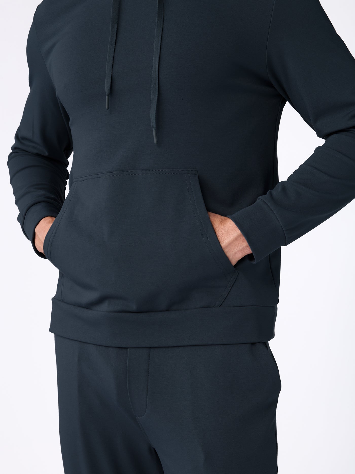 A man models the Cozy Earth Men's Brushed Bamboo Hoodie & Jogger Set in a dark color, standing with both hands in the hoodie’s front pocket, shown from shoulders to mid-thigh against a plain light background. 