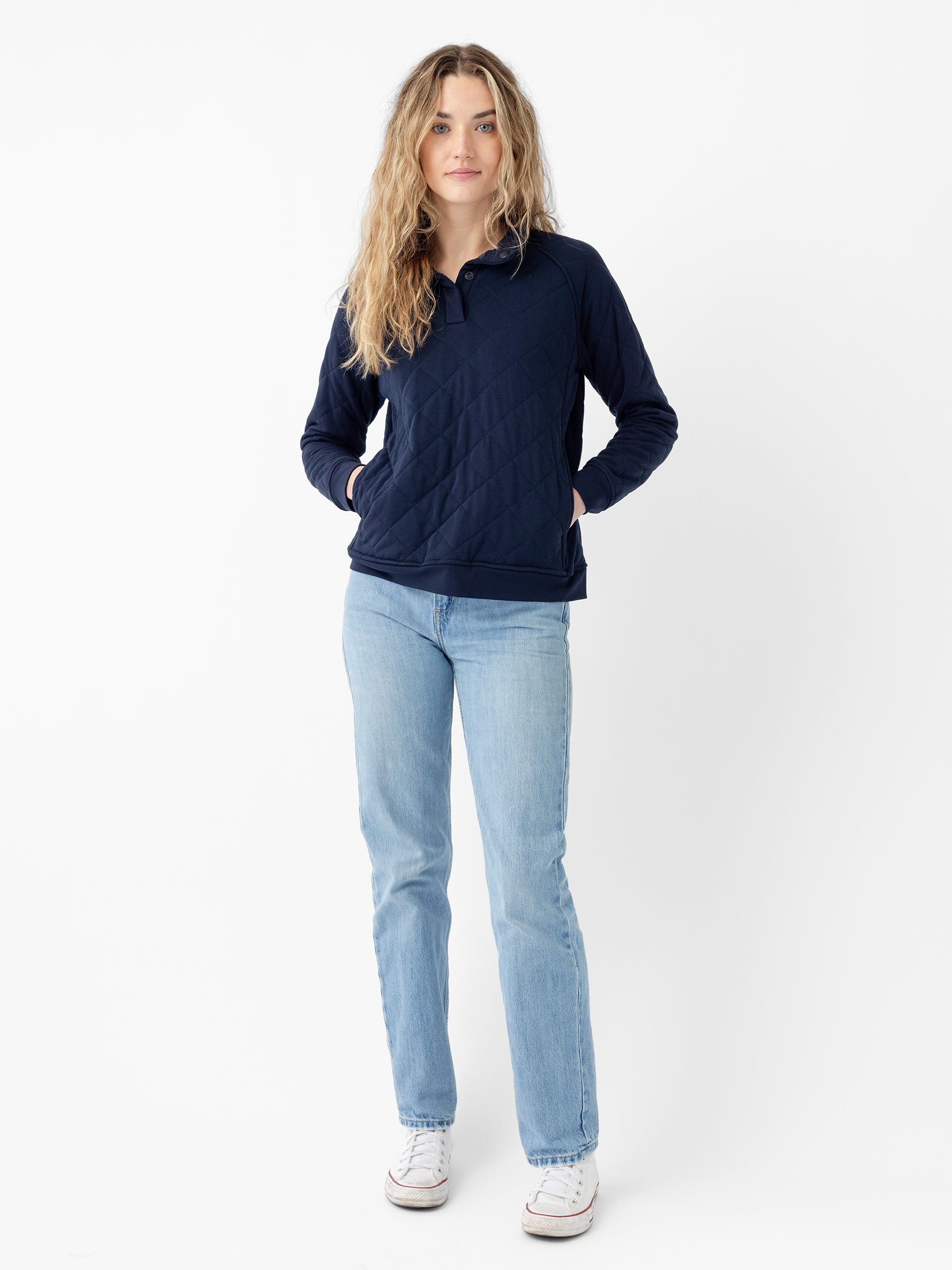 A person with long, wavy hair is wearing a Cozy Earth Women's Ultra-Soft Bamboo Quilted Snap Pullover in navy blue, light blue jeans, and white sneakers. They are standing against a plain white background with their hands in their pullover pockets, looking directly at the camera. 