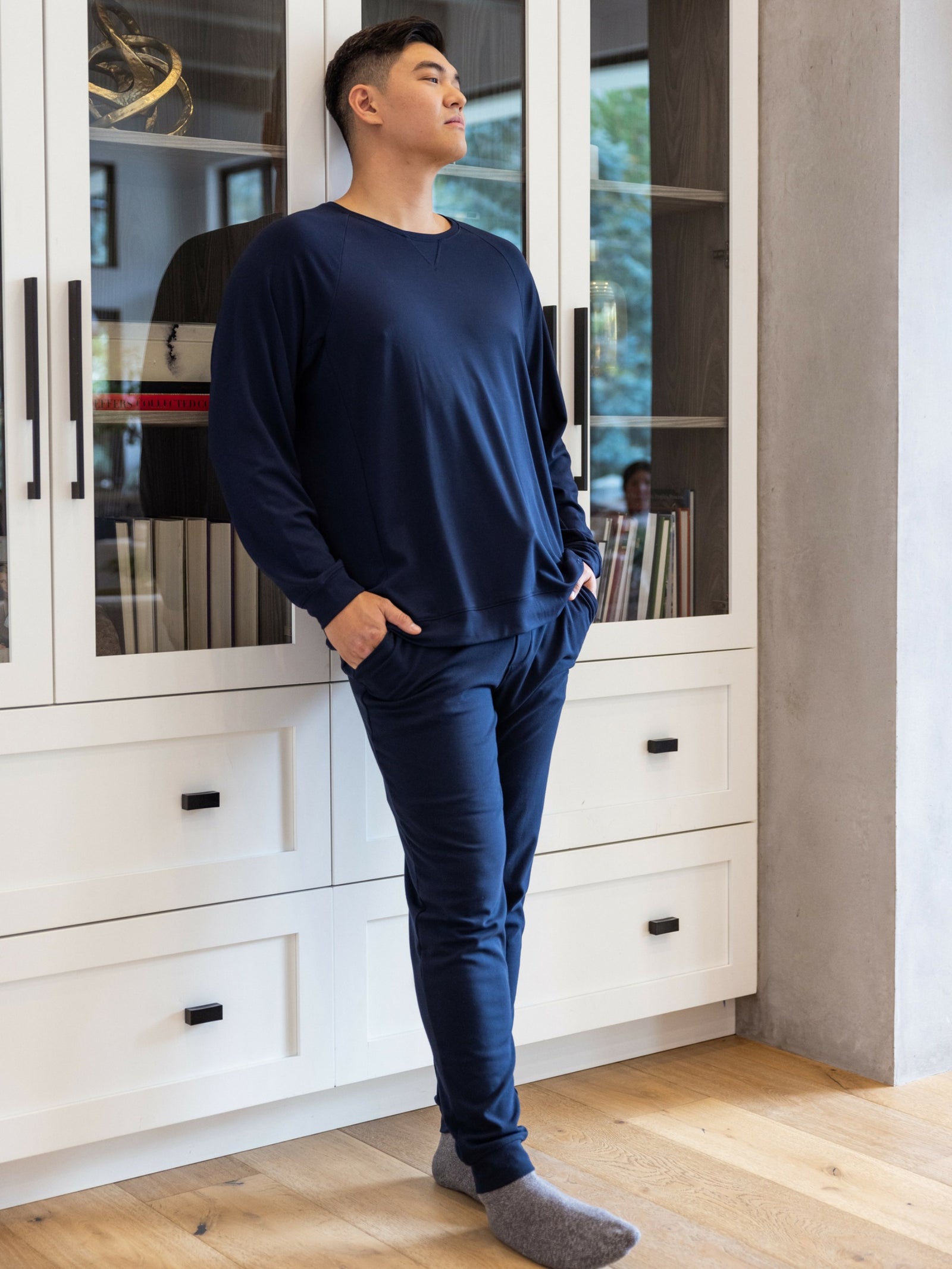 A person in a Cozy Earth Men's Bamboo Jogger Set stands with hands in pockets before a white cabinet, wearing gray socks and gazing upward. The scene includes books and a window revealing trees. 