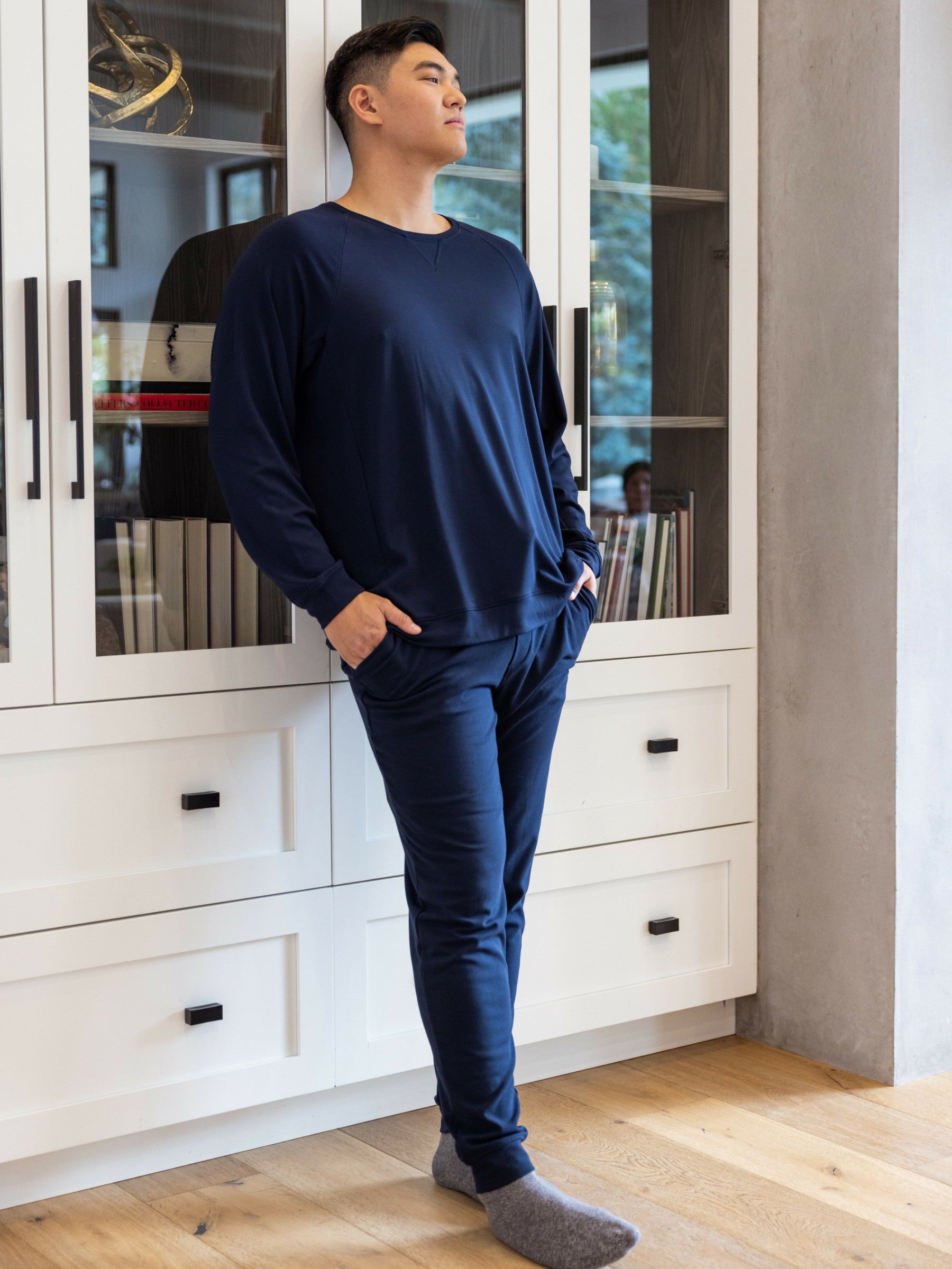 A person in a Cozy Earth Men's Bamboo Jogger Set stands with hands in pockets before a white cabinet, wearing gray socks and gazing upward. The scene includes books and a window revealing trees. |Color:Navy