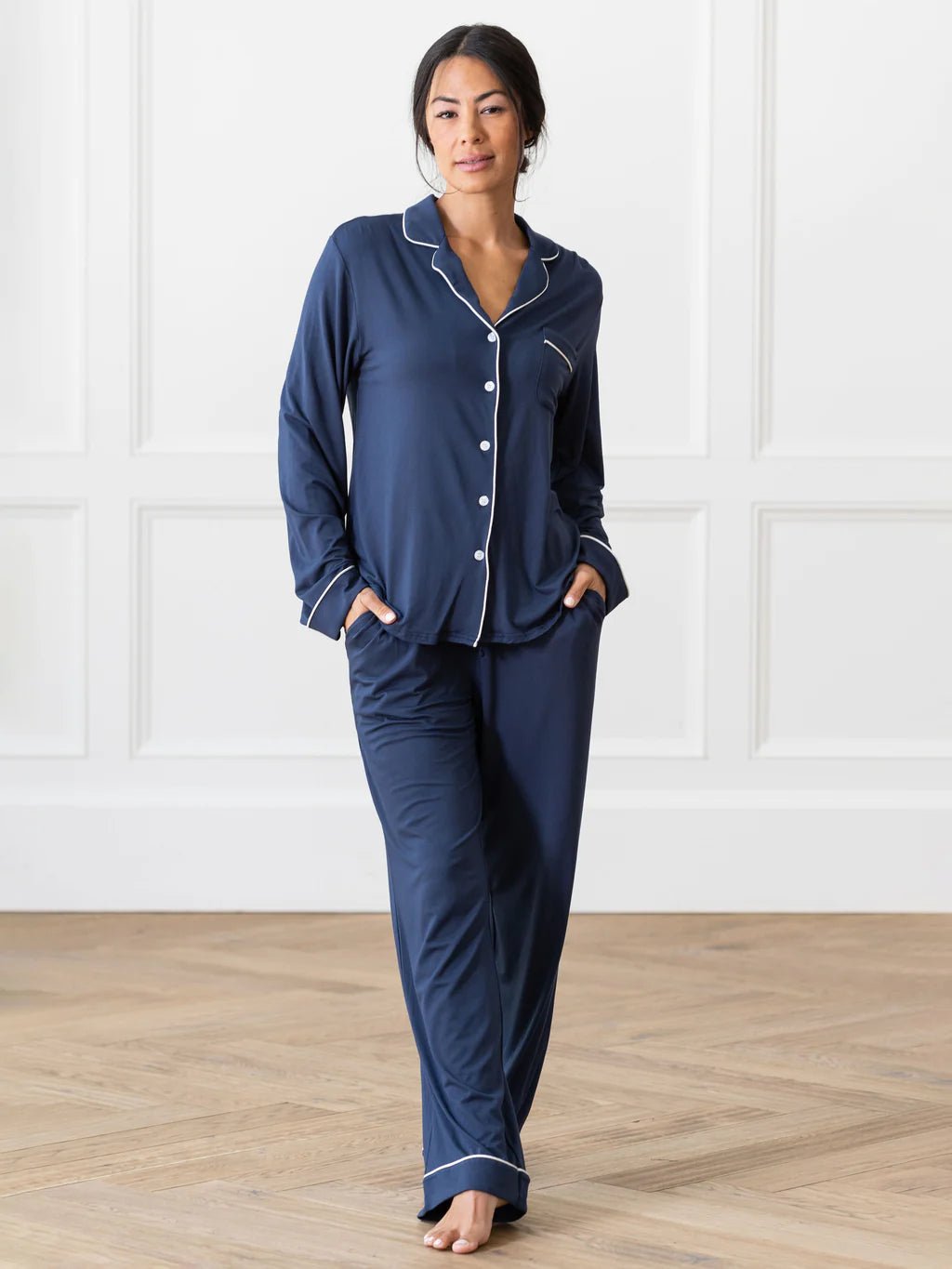 A person stands barefoot on a wooden floor, dressed in the Women's Bedtime Bundle from Cozy Earth. The outfit features navy blue long-sleeved button-up pajamas with white trim and matching long pants. The scene is set against a backdrop of white paneled walls. 