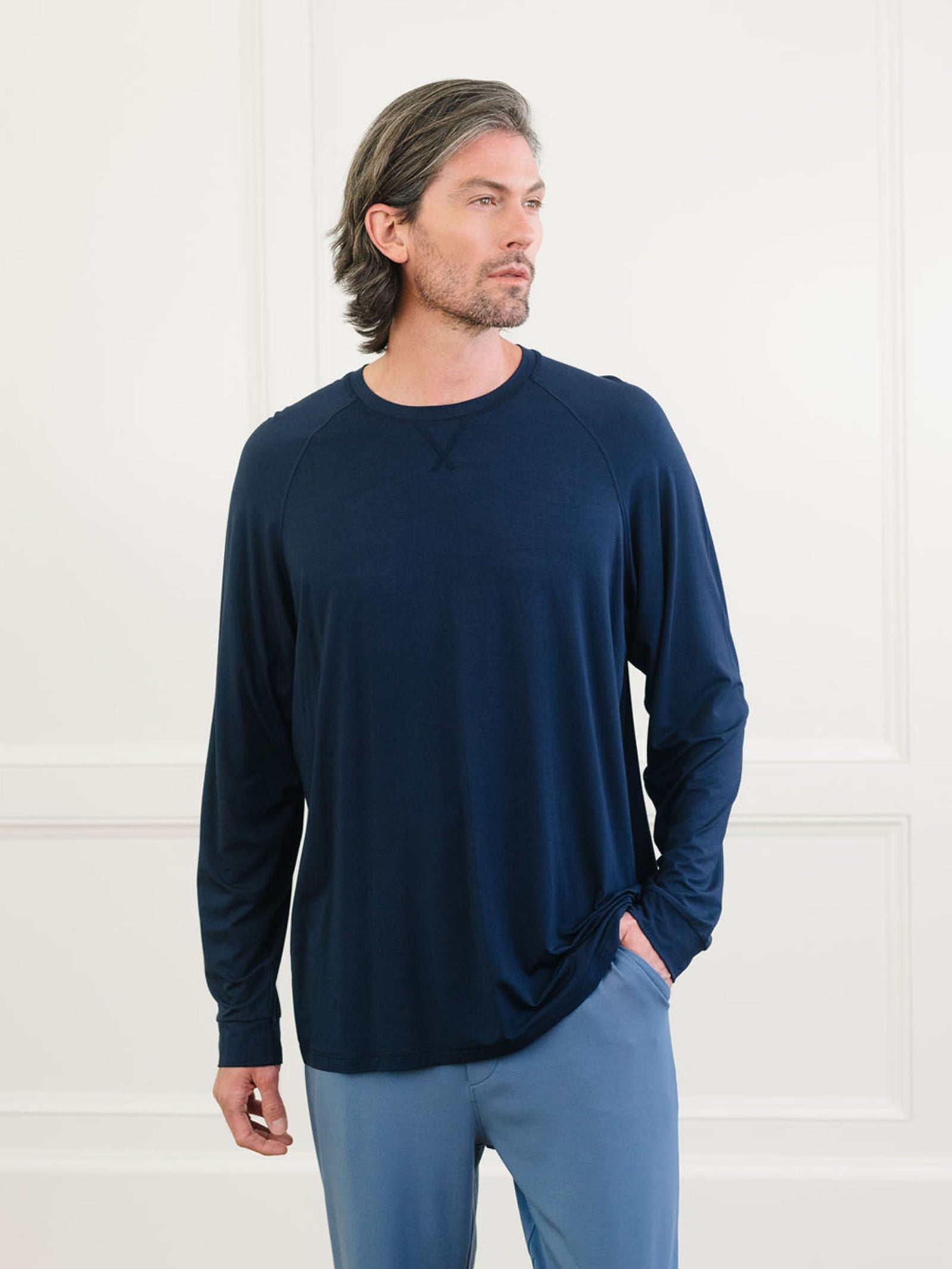 A man with shoulder-length hair and a beard is wearing the Cozy Earth Men's Stretch-Knit Long Sleeve Bamboo Pajama Jogger Set in navy blue. He is standing indoors against a light-colored background with wainscoting, looking to his left with one hand in his pocket. 