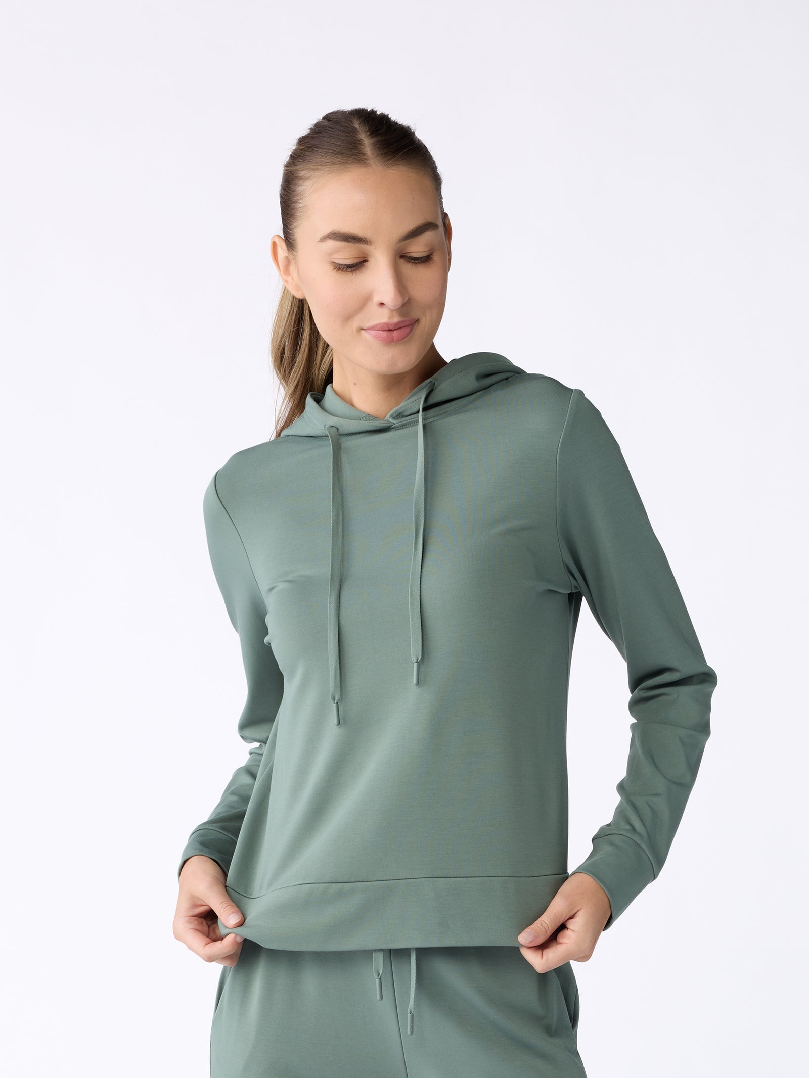 A person wearing a Women's Ultra-Soft Bamboo Hoodie from Cozy Earth stands against a plain white background. They have long hair tied back and are looking downwards, lightly touching the hem of the hoodie with both hands. The hoodie features drawstrings and has a relaxed fit. 