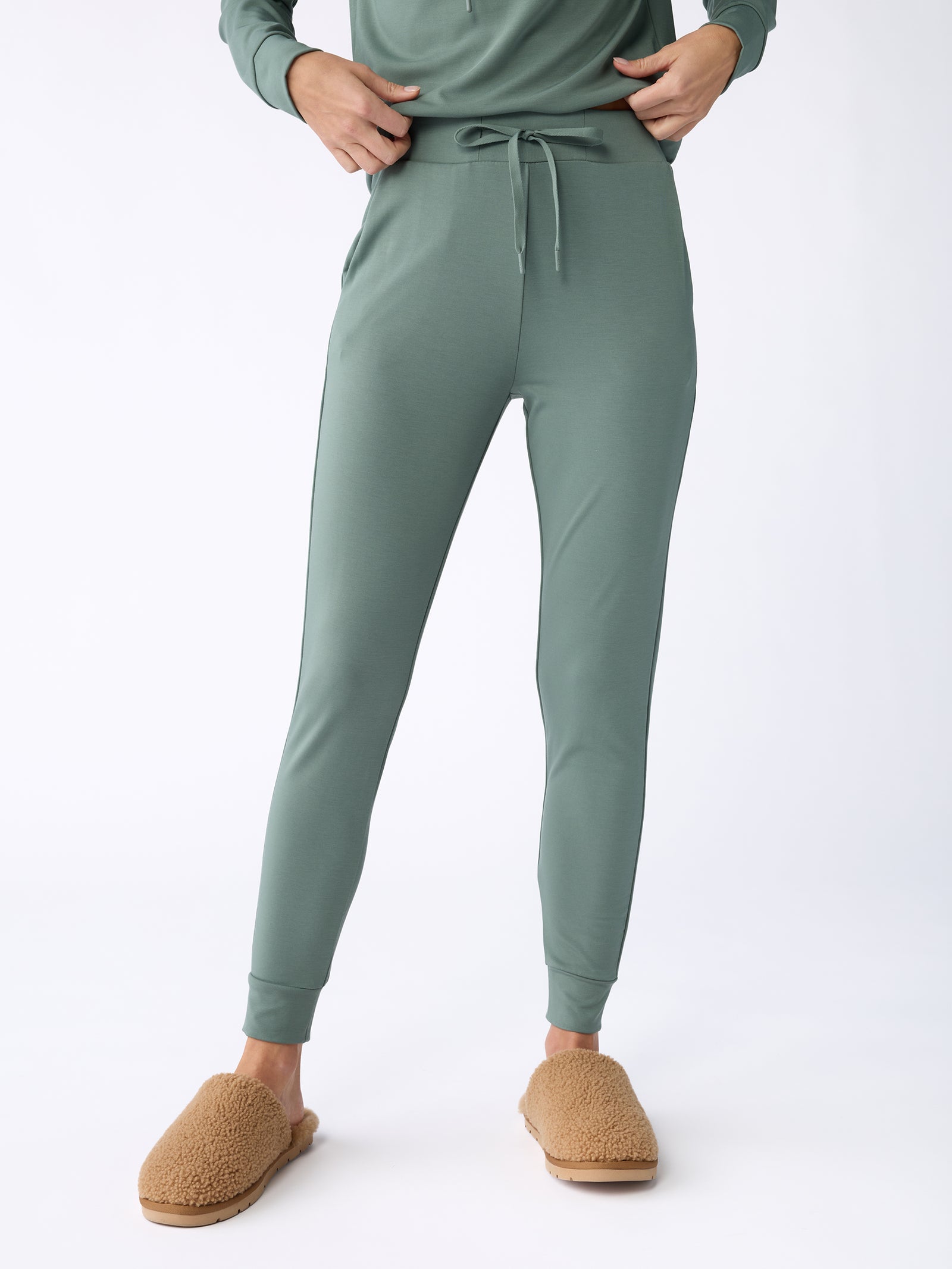 A person is standing while wearing the Women's Bamboo Jogger Pant in sage green from Cozy Earth, paired with a matching top. Their hands rest in the pants pockets, and they have on tan, fuzzy slippers. The background is plain white. 