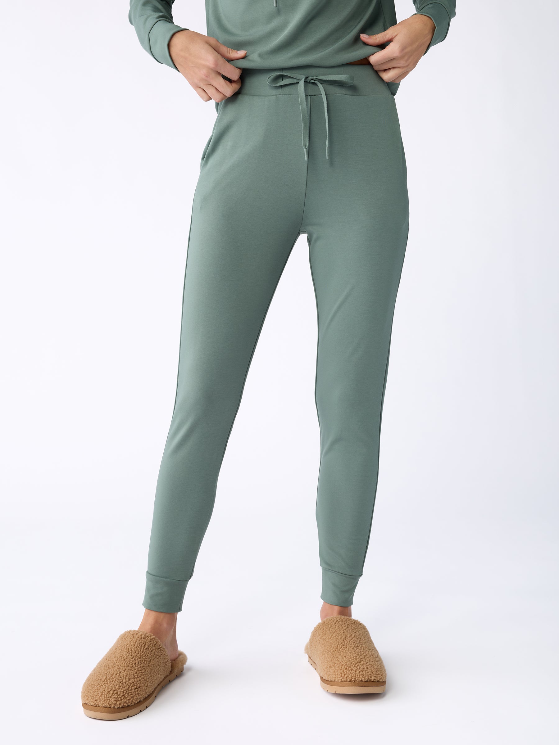 A person is standing while wearing the Women's Bamboo Jogger Pant in sage green from Cozy Earth, paired with a matching top. Their hands rest in the pants pockets, and they have on tan, fuzzy slippers. The background is plain white. |Color:Oakmoss