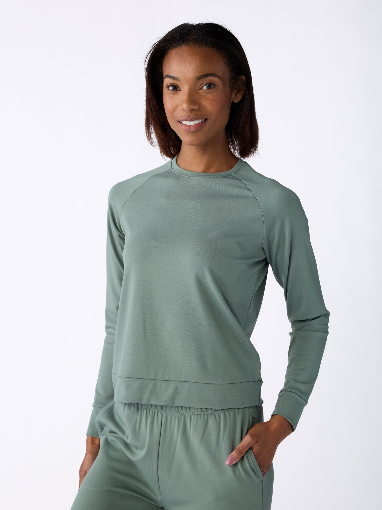 A person wearing the Women's Ultra-Soft Bamboo Pullover Crew from Cozy Earth stands against a plain white background. They are smiling and have one hand in a pocket, exuding a relaxed and casual vibe, with their outfit featuring a matching long-sleeve top and pants. 