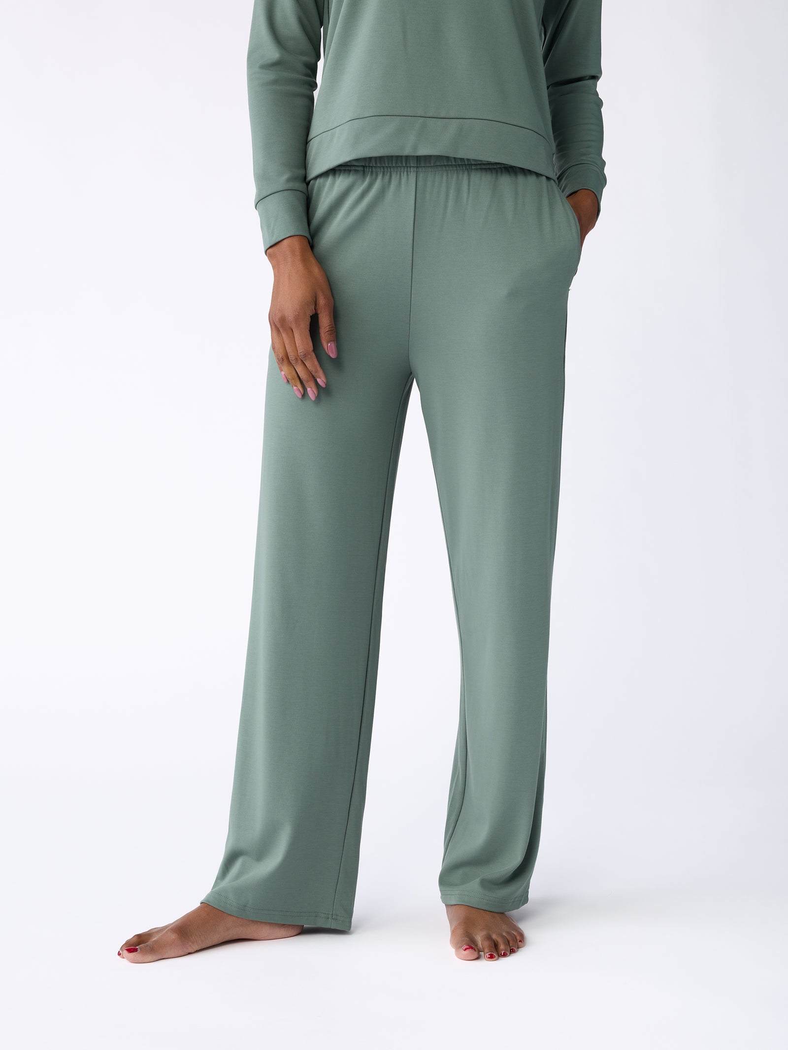 A person is standing barefoot against a plain white background, wearing the Women's Ultra-Soft Bamboo Wide Leg Pull On Pant from Cozy Earth in sage green. The loungewear includes a long-sleeved top and relaxed-fit pants, with one hand casually placed in the pocket. 