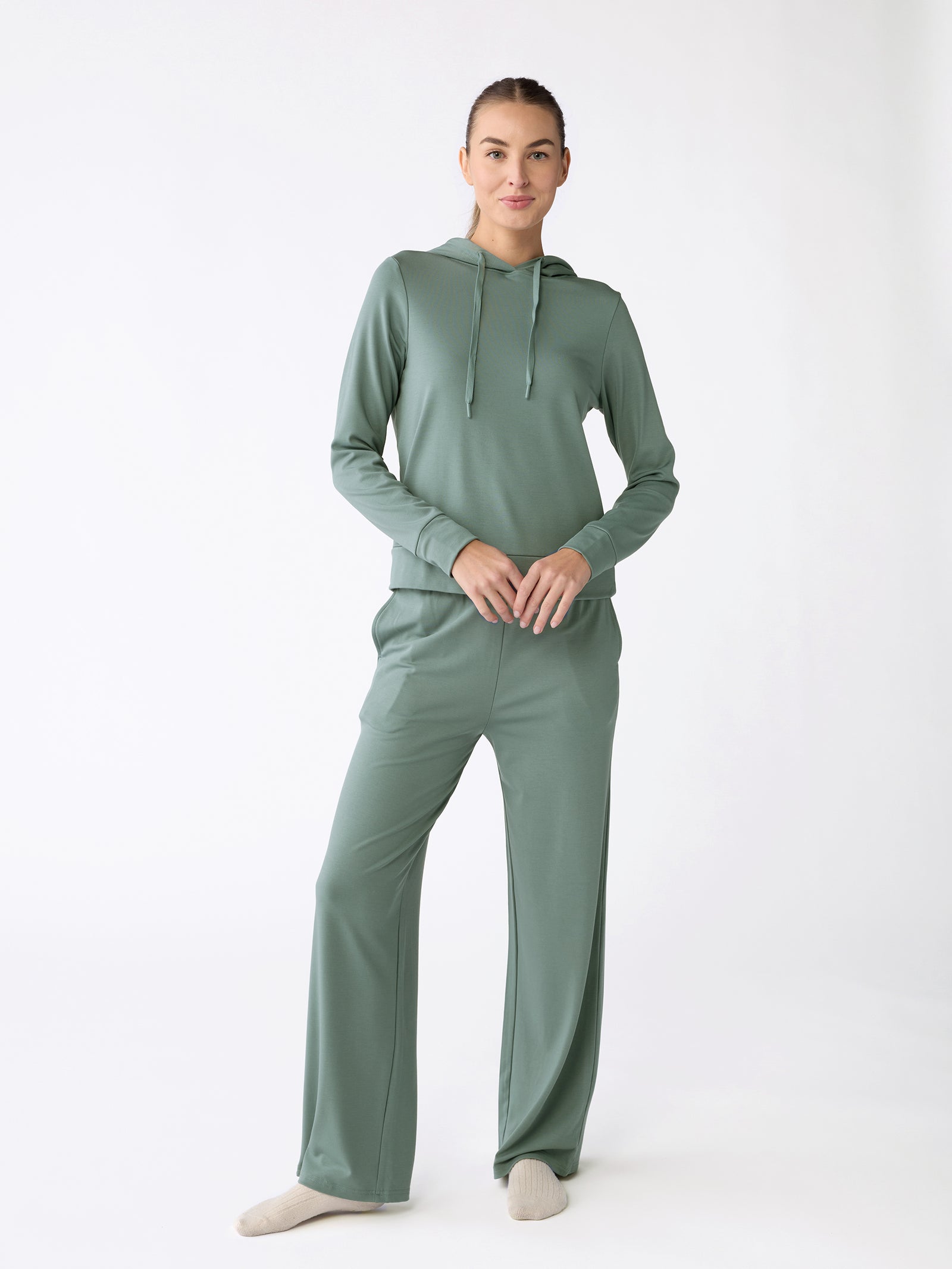 A person models the Women's Ultra-Soft Bamboo Wide Leg Pull On Pant & Hoodie Set in light green by Cozy Earth, adopting a relaxed pose against a plain white background while wearing beige socks. 