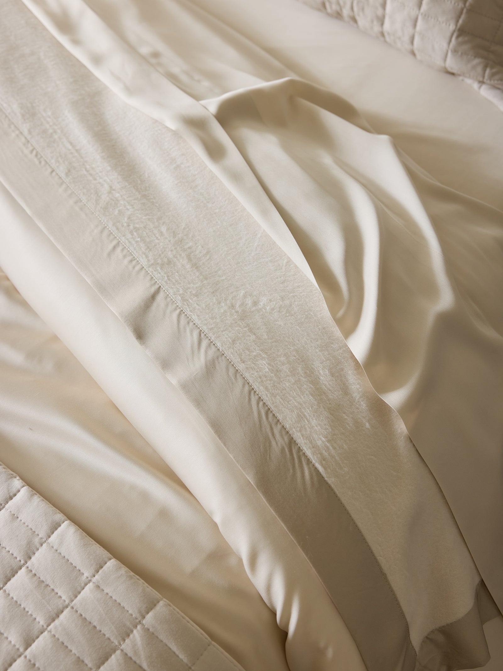 A close-up of a neatly made bed featuring ivory-colored satin sheets and a quilted Bamboo Blanket by Cozy Earth. The satin sheets reflect light and appear smooth with subtle wrinkles, while the Bamboo Blanket adds an extra layer of luxurious comfort. 