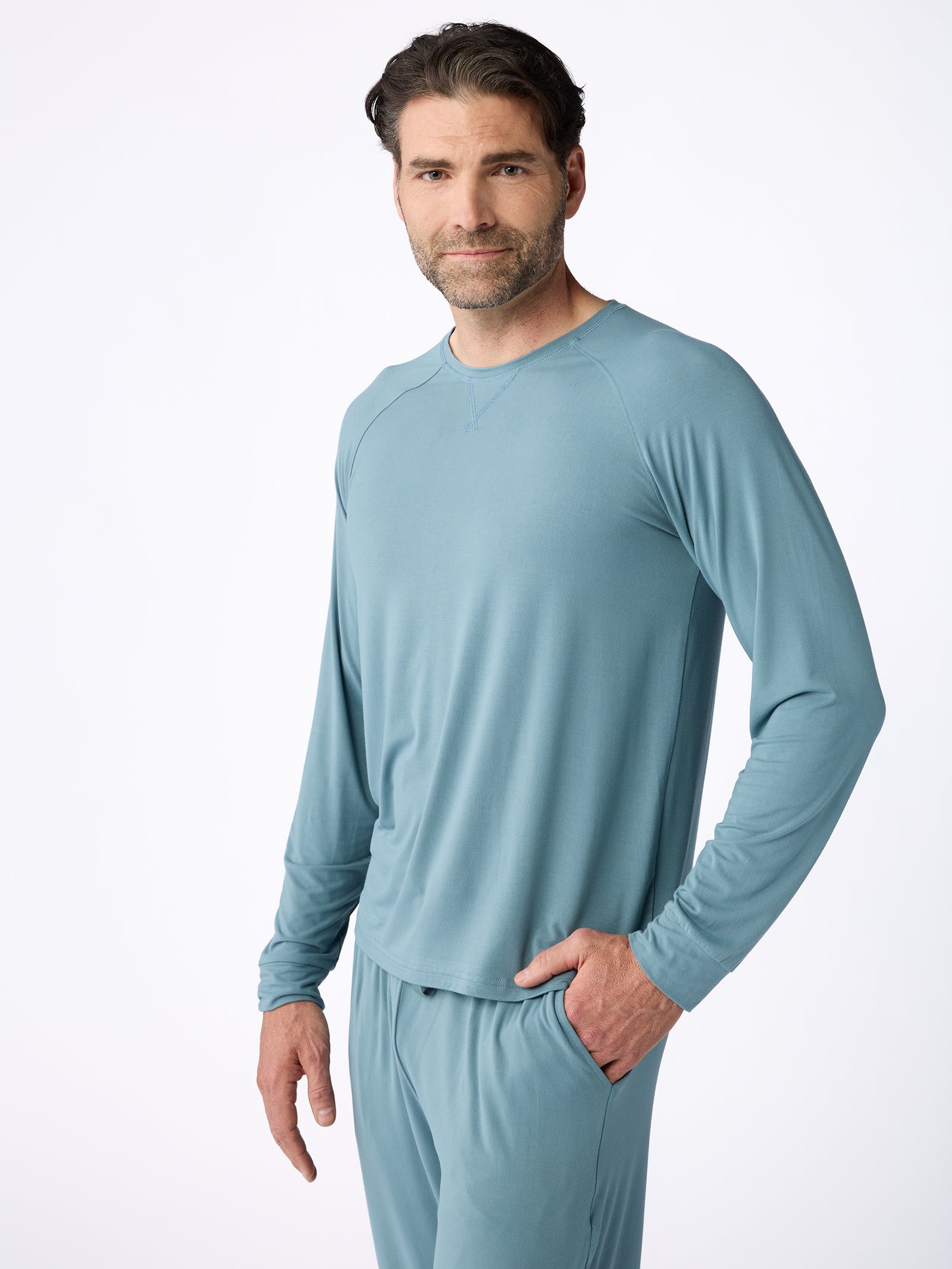 A man with short dark hair and a beard is dressed in a light blue Men's Stretch-Knit Bamboo Long Sleeve by Cozy Earth and matching pants. He stands against a plain white background with his right hand in his pocket, gazing at the camera with a neutral expression. 