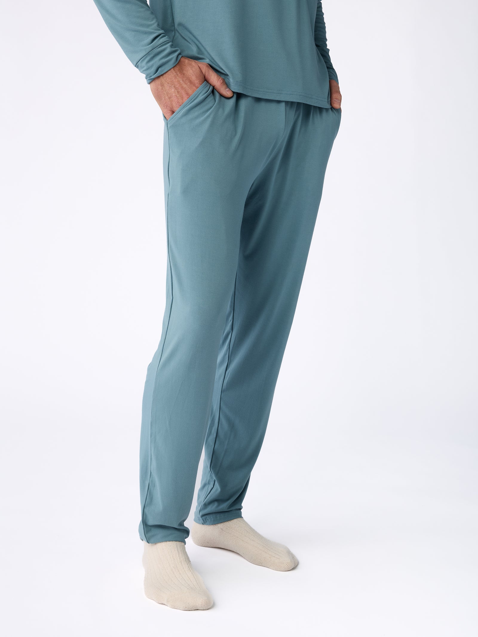 A person is wearing the Men's Stretch-Knit Bamboo Pajama Pant in teal by Cozy Earth, along with a matching long-sleeve top. The pants have a relaxed fit, and the person completes the look with beige socks, set against a plain white background. 