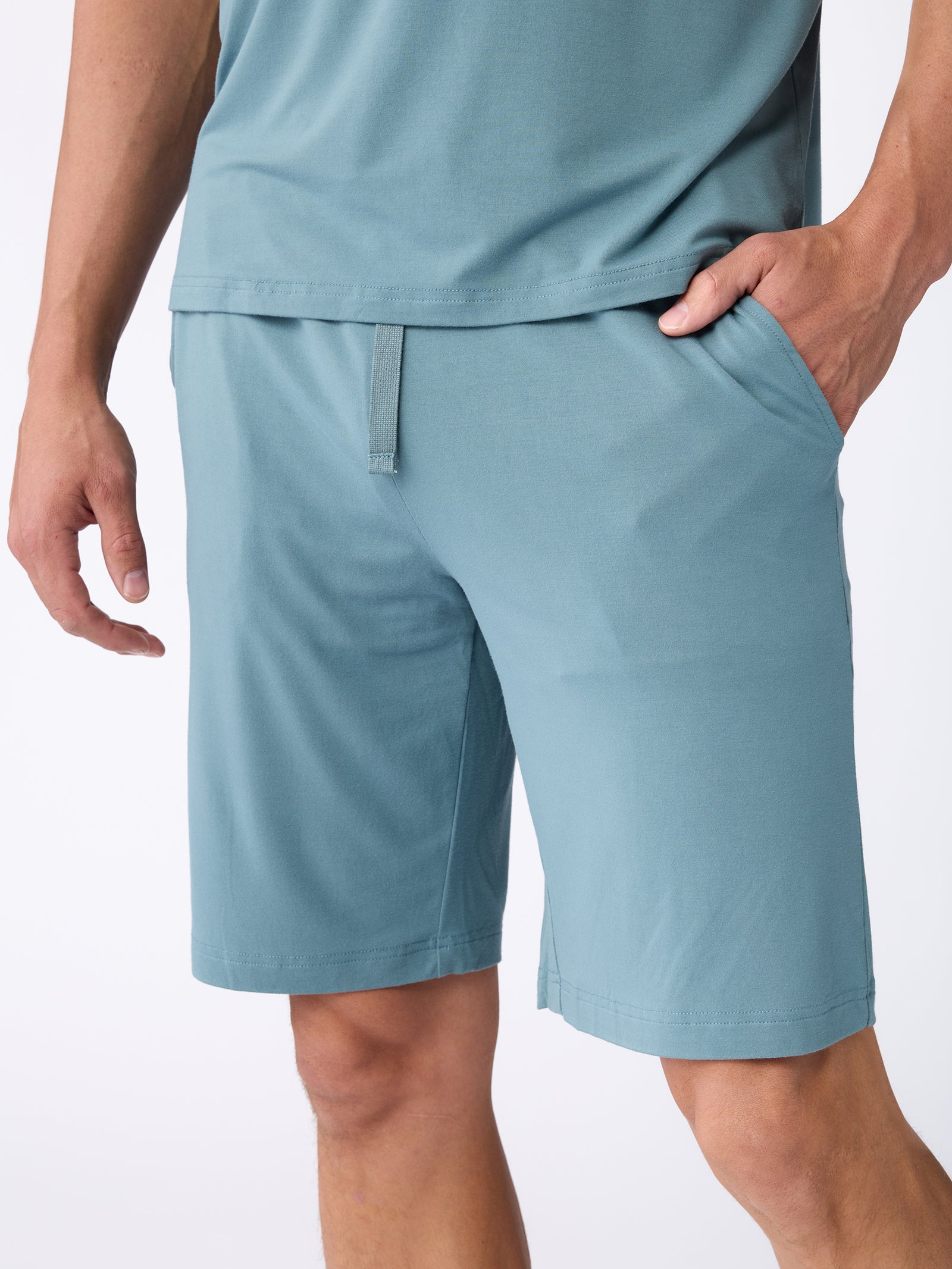 The individual is dressed in a teal ensemble from Cozy Earth, featuring a short-sleeve shirt paired with the Men's Stretch-Knit Bamboo Pajama Short. One hand is casually placed in the pocket of the shorts against a plain white backdrop. 